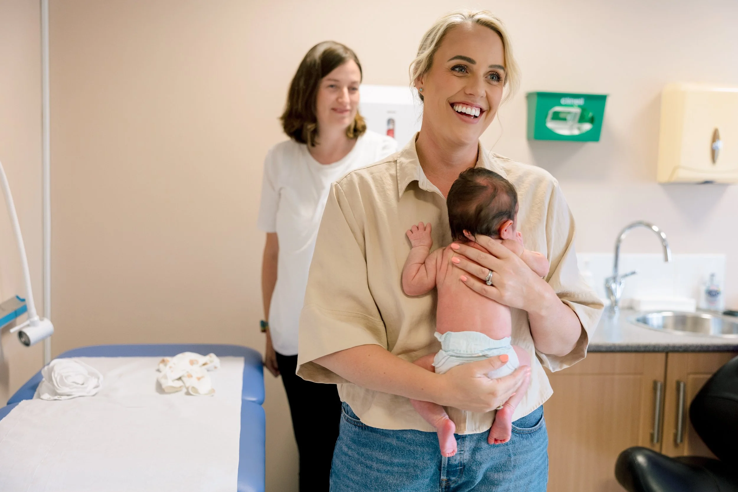 A smiling woman holding a newborn baby in a hospital room, with a nurse or healthcare worker standing behind her. There is a hospital bed and medical equipment visible.