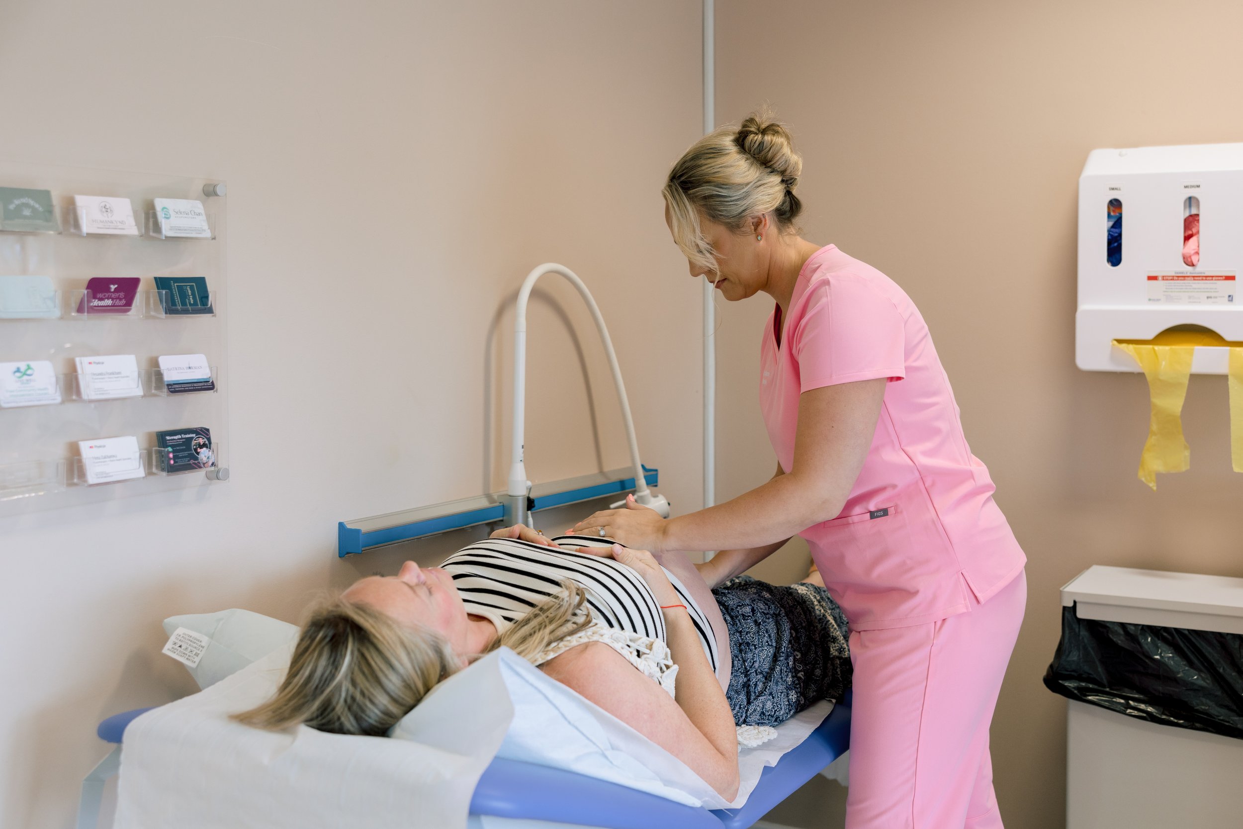 A nurse in pink scrubs attending to a pregnant woman lying in a hospital bed, with medical equipment and supplies on the wall behind them.