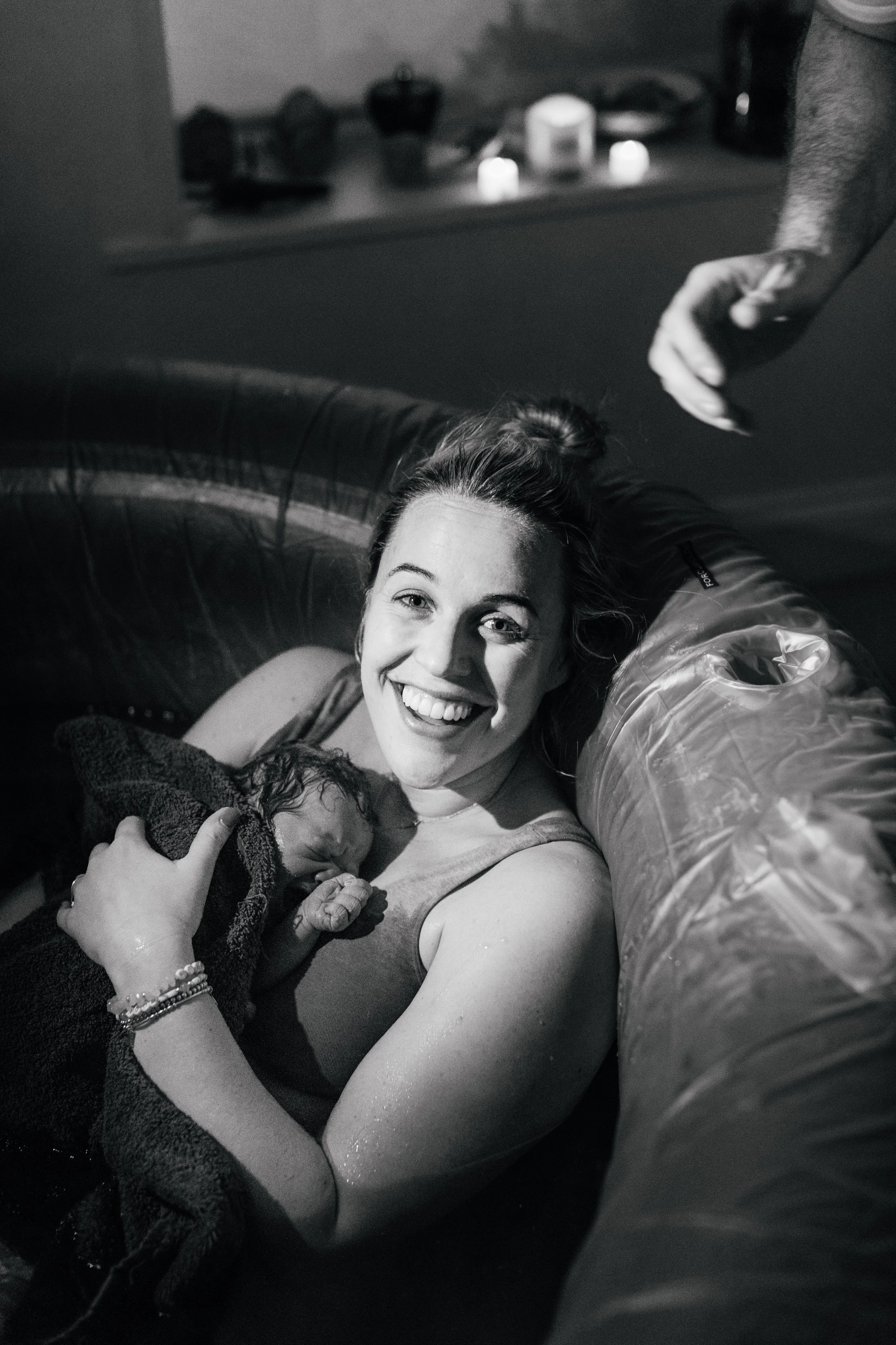 A woman with a smile smiling at the camera, holding a newborn baby, possibly a girl, wrapped in a dark blanket, lying on her chest. The woman is wearing a sleeveless top, and they are in an indoor setting. An arm with an outstretched hand is visible in the upper right corner of the image.