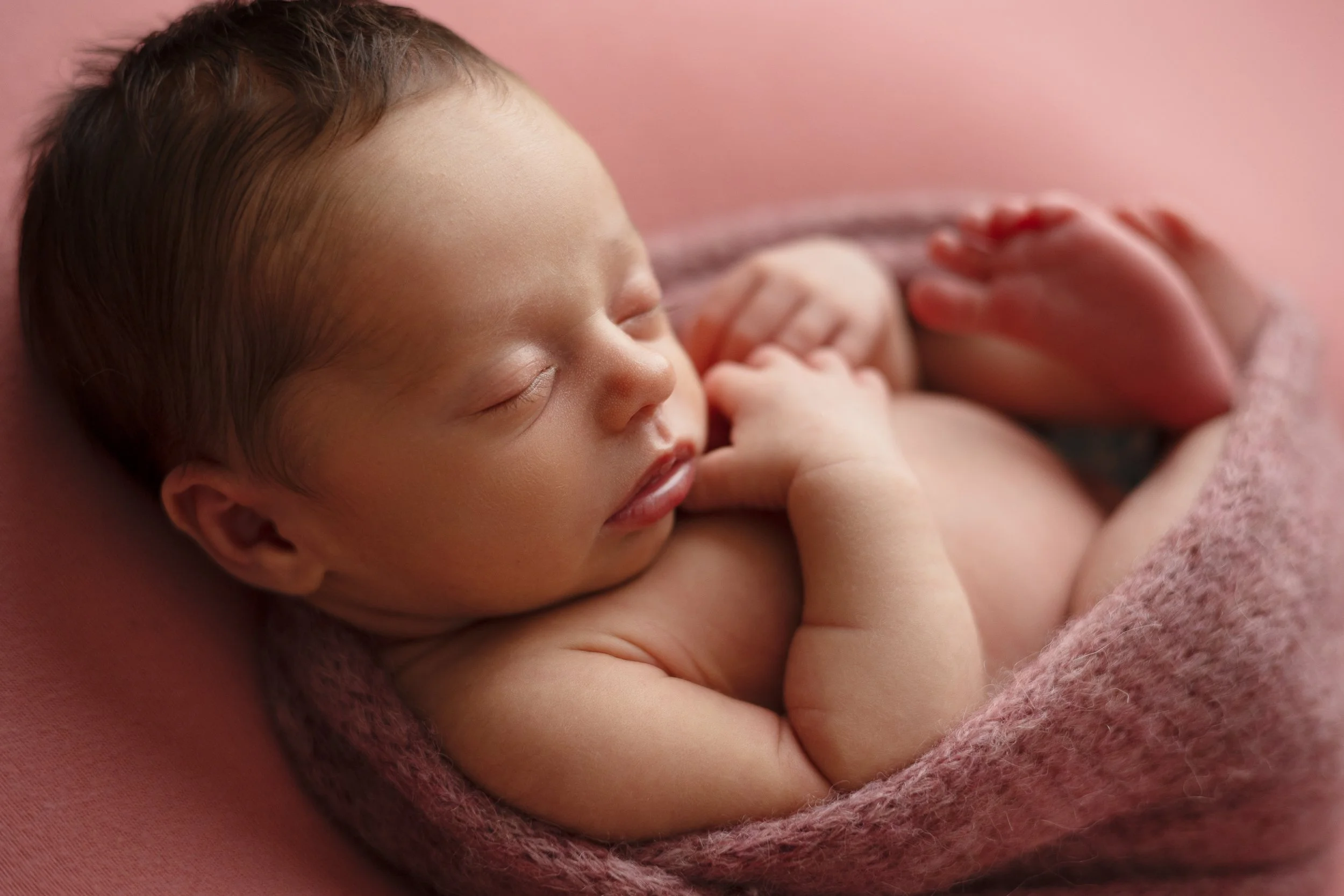 Newborn baby girl wrapped in a soft pink hand knit wrap on a pink backdrop.