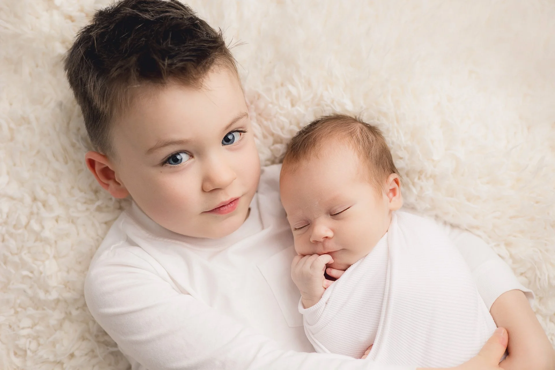 newborn baby wrapped in a white stretchy wrap being help by his older brother on a cream blanket baby is sleeping older boy is looking at the camera