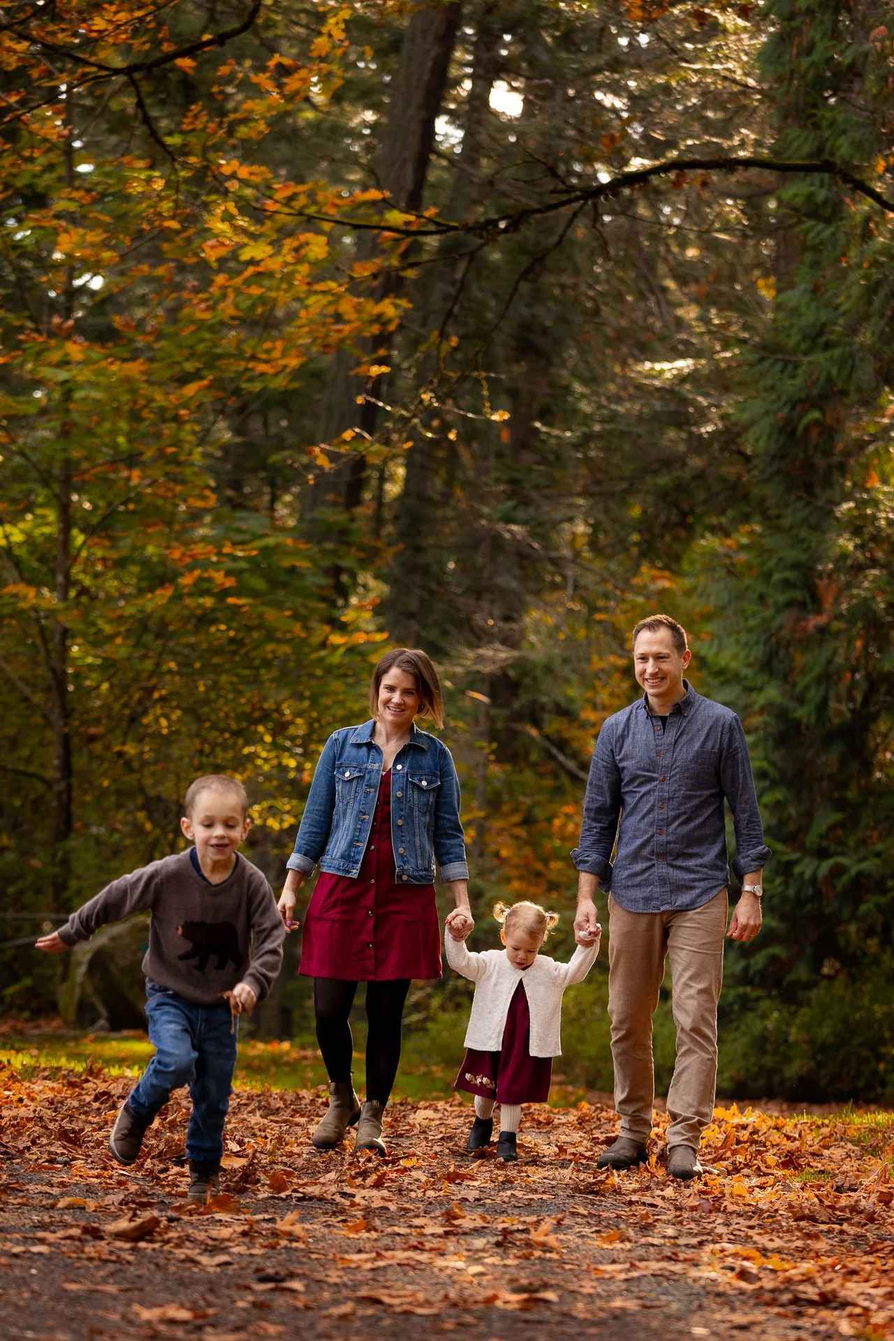 Family of four photographed at mount doug park in Victoria BC holding hand and walking through the fall leaves. Young boy is running away from family laughing.  