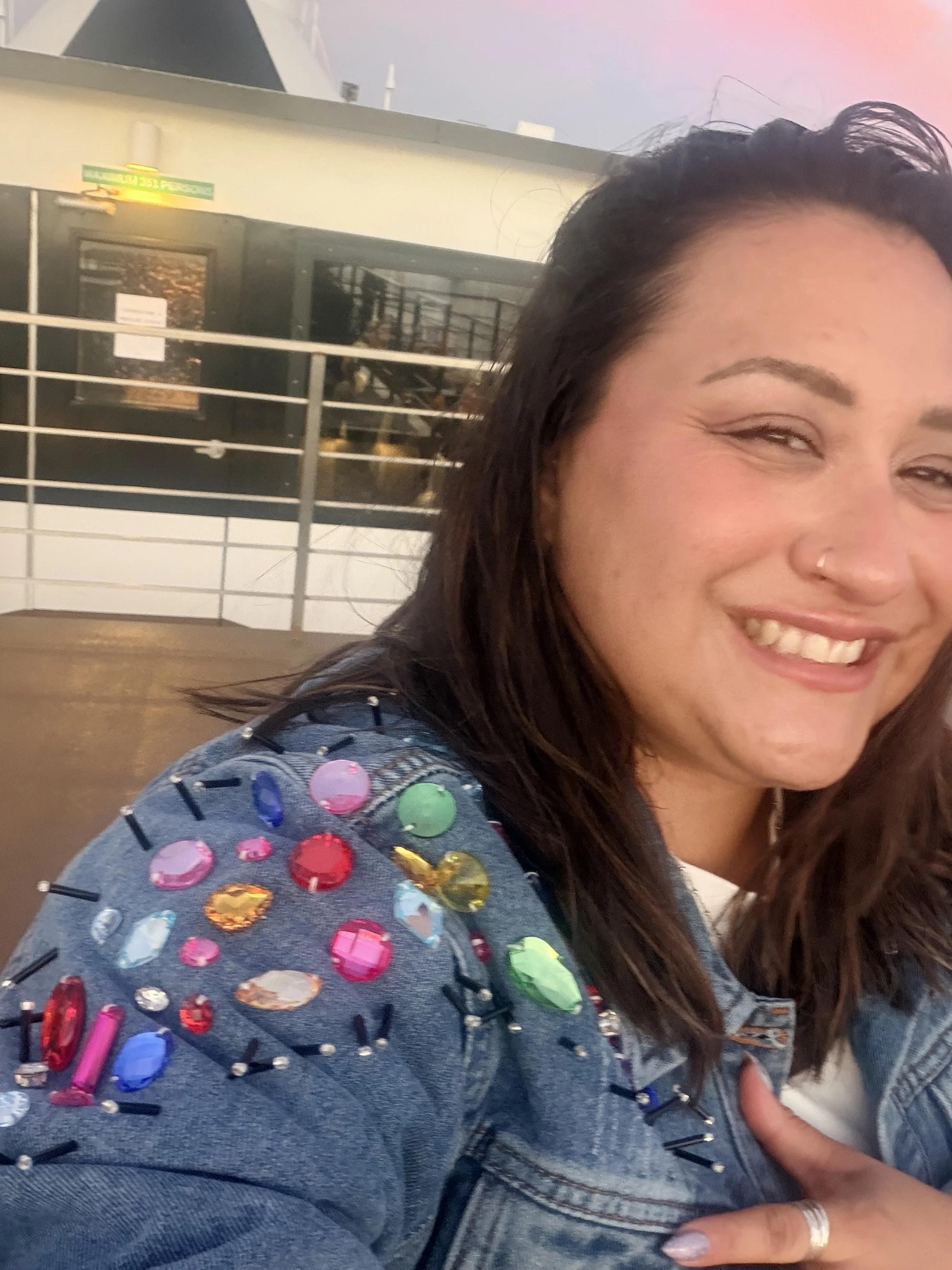 A smiling woman with black wavy hair, wearing a denim jacket decorated with colorful gemstones, standing outdoors against a building and metal railing.