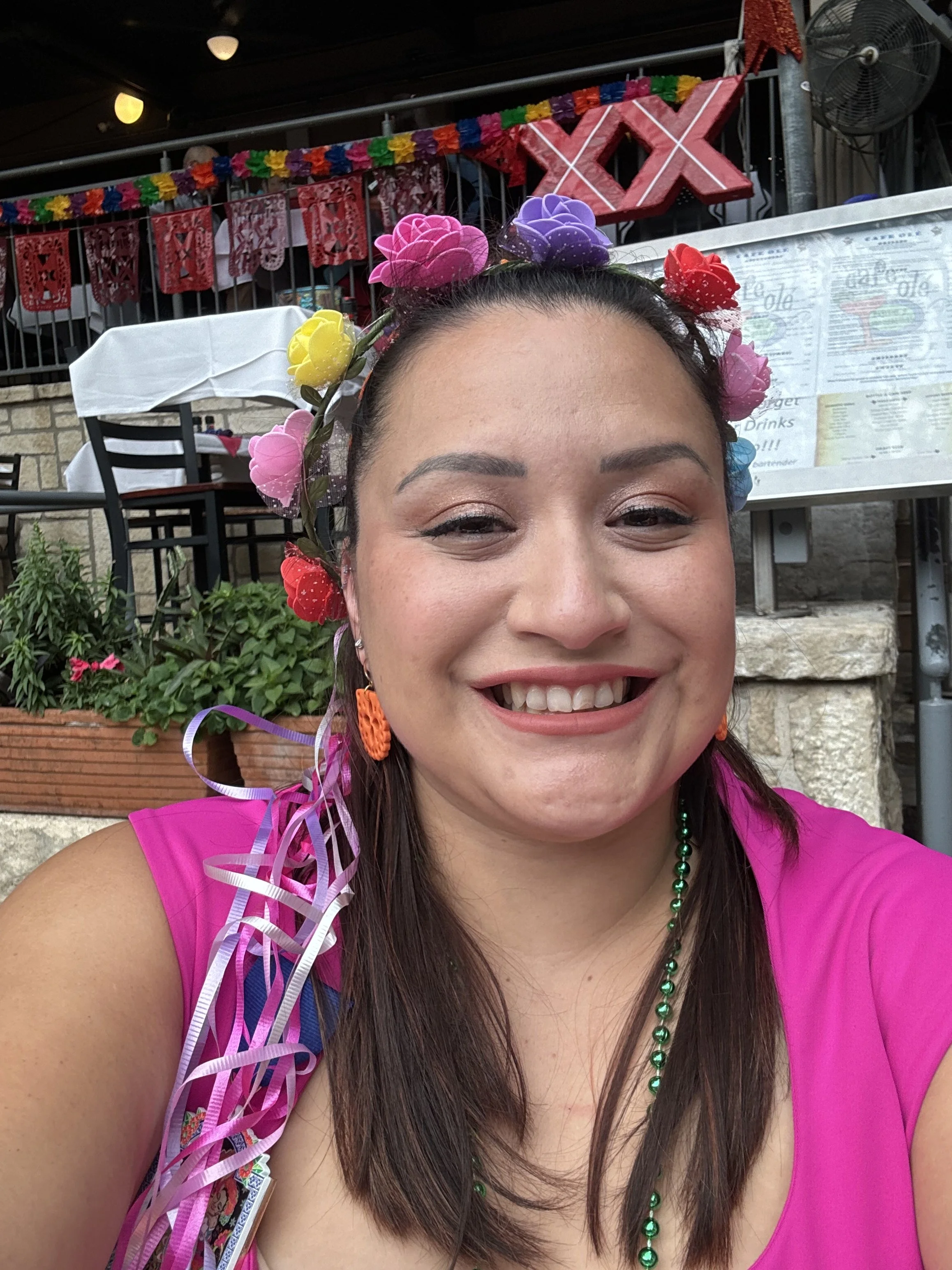 Close-up of a woman with a flower crown, smiling, at a celebration with decorative banners and signs reading 'XIX' in the background.