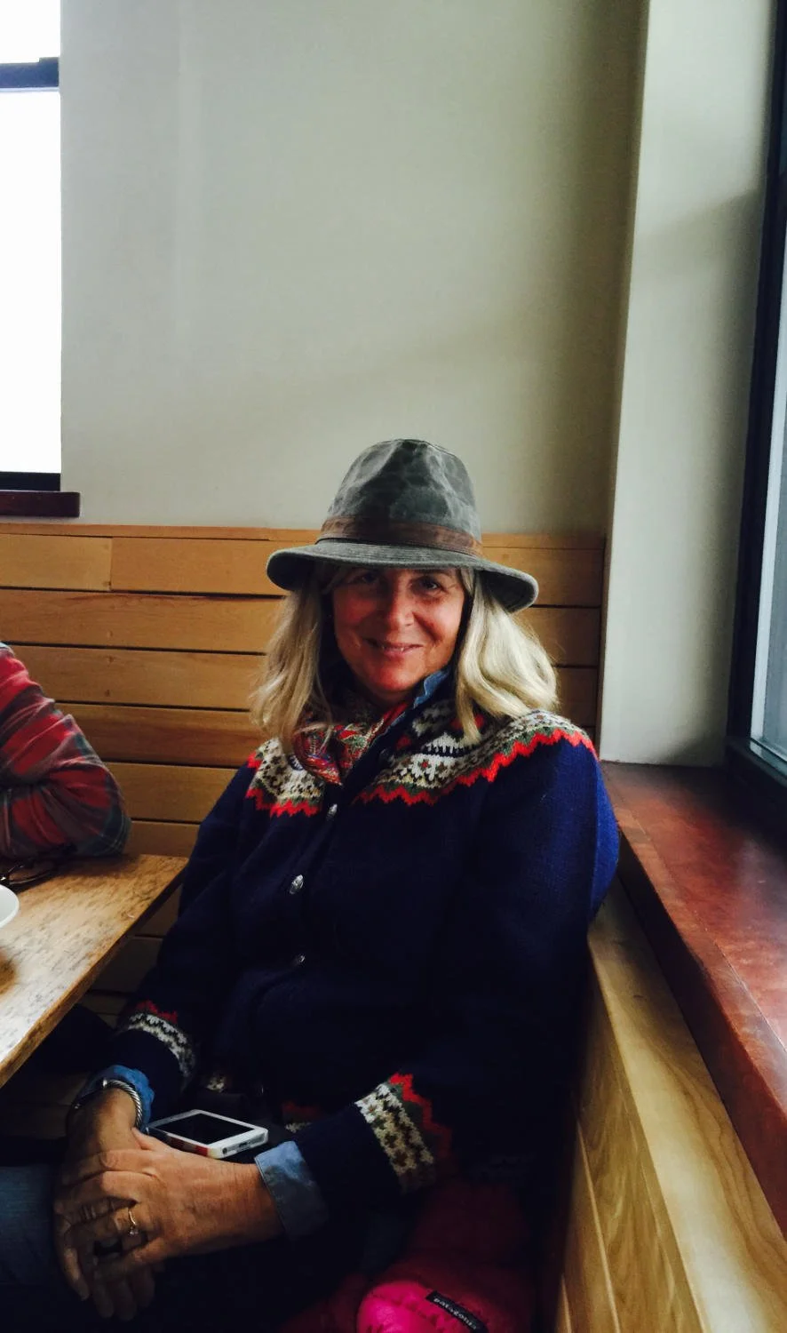 Woman with blonde hair wearing a gray hat and a dark sweater with colorful patterns, sitting at a wooden table in a cozy indoor setting.