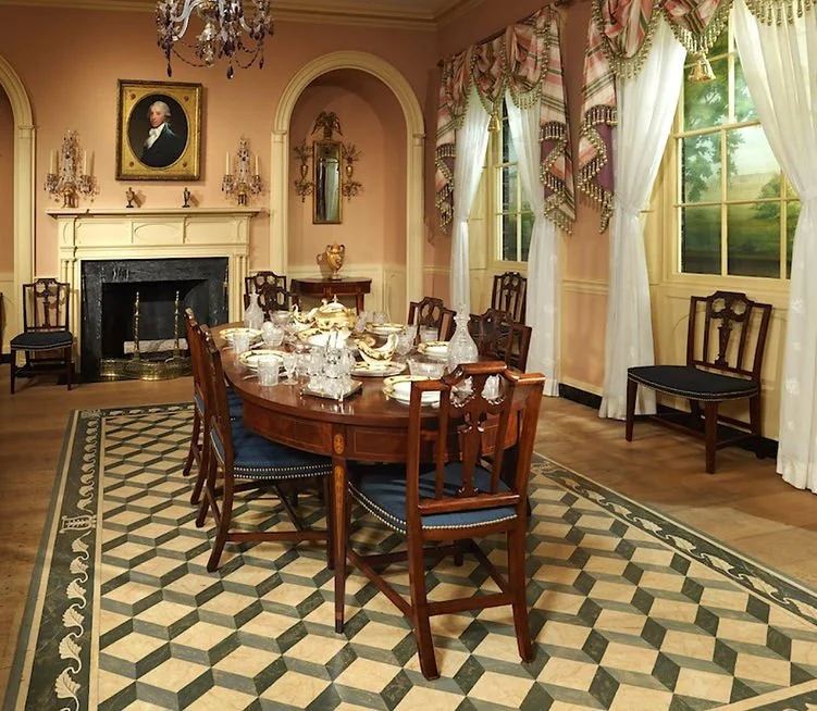 Elegant dining room with a large wooden table set for a meal, surrounded by wooden chairs with blue cushions, a fireplace with a portrait above, and grand windows with ornate curtains, in a classic style.