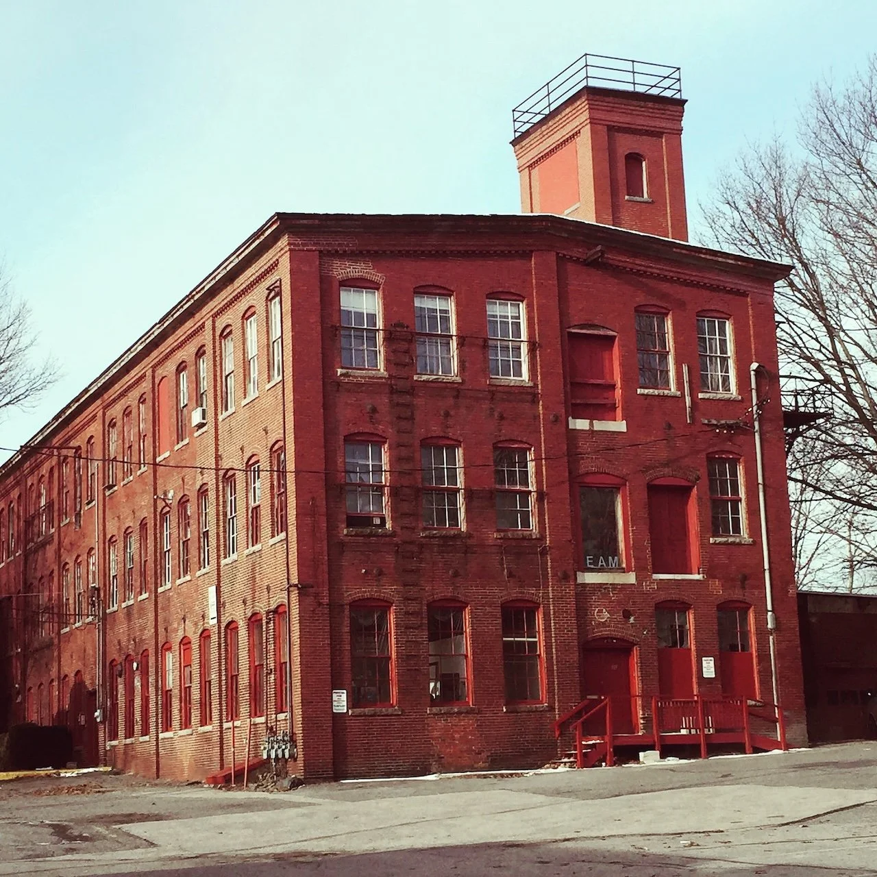 A red brick building with multiple floors, tall windows, and a small tower on top, with some trees and power lines nearby.