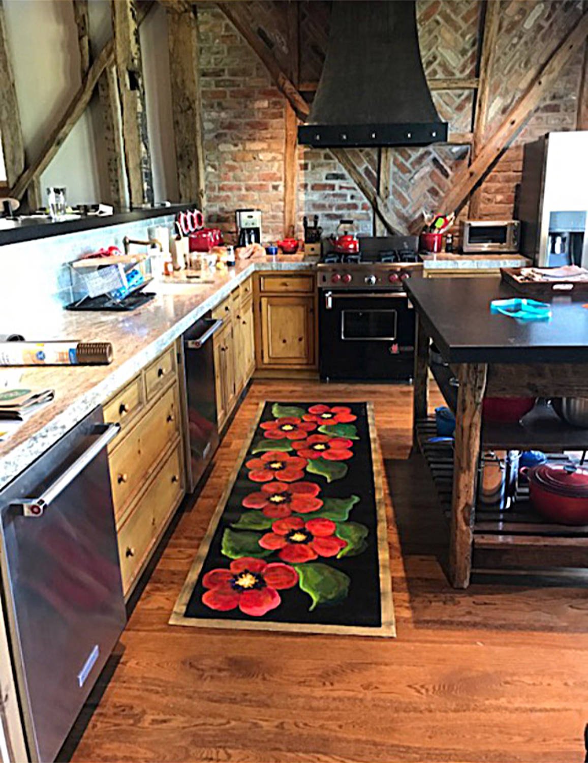 A rustic kitchen with wood cabinets, a black stove, and brick walls. There is a floral runner rug on the wooden floor, and various kitchen appliances and utensils on the countertops.
