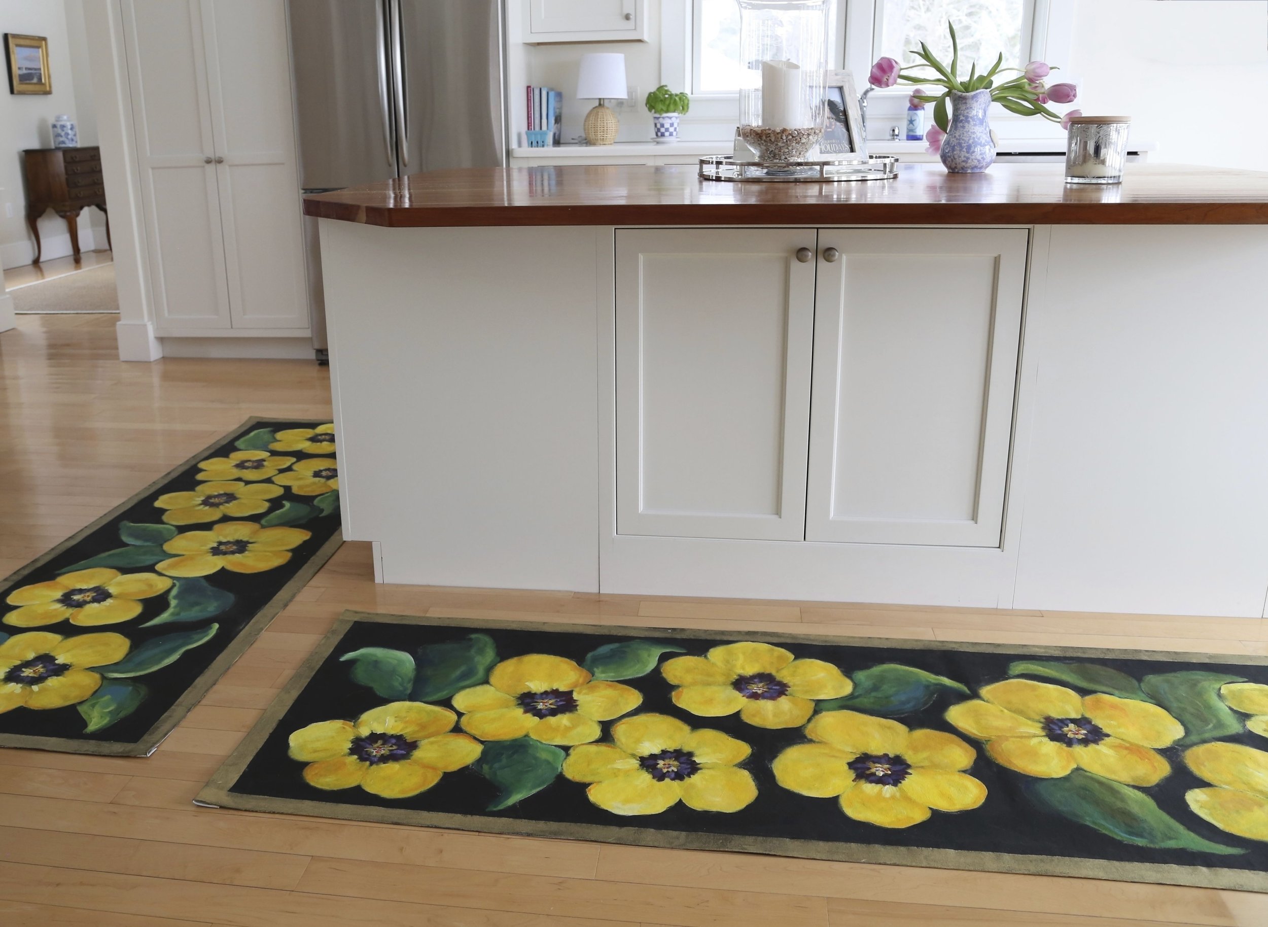 Colorful floral rugs with yellow flowers and green leaves on a wooden floor in a kitchen. The kitchen has white cabinets, a brown wooden countertop, and a window with sunlight.