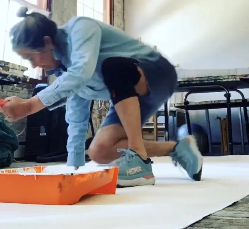Person bending down on one knee, touching things on the floor in a room with large windows and tables.
