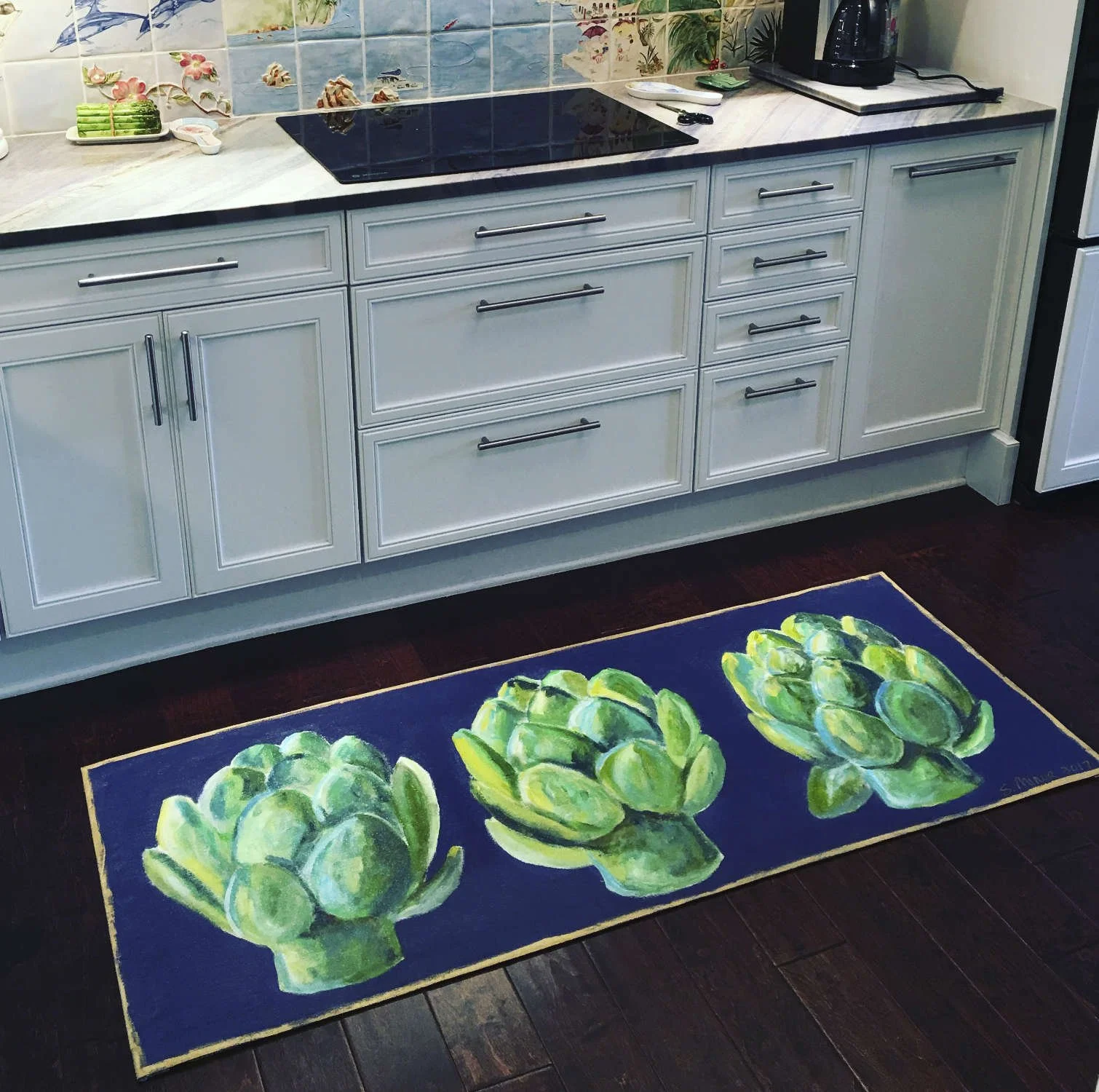 A colorful kitchen with white cabinets, a black stovetop, and a backsplash featuring a beach scene with seashells and tropical plants. On the dark wood floor, there's a rectangular rug with painted green artichokes on a dark blue background.