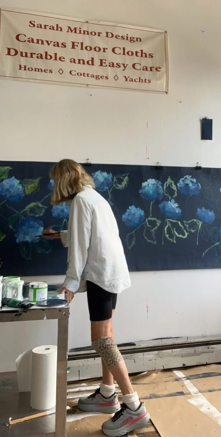 A woman painting a mural of blue hydrangea flowers on a dark background.