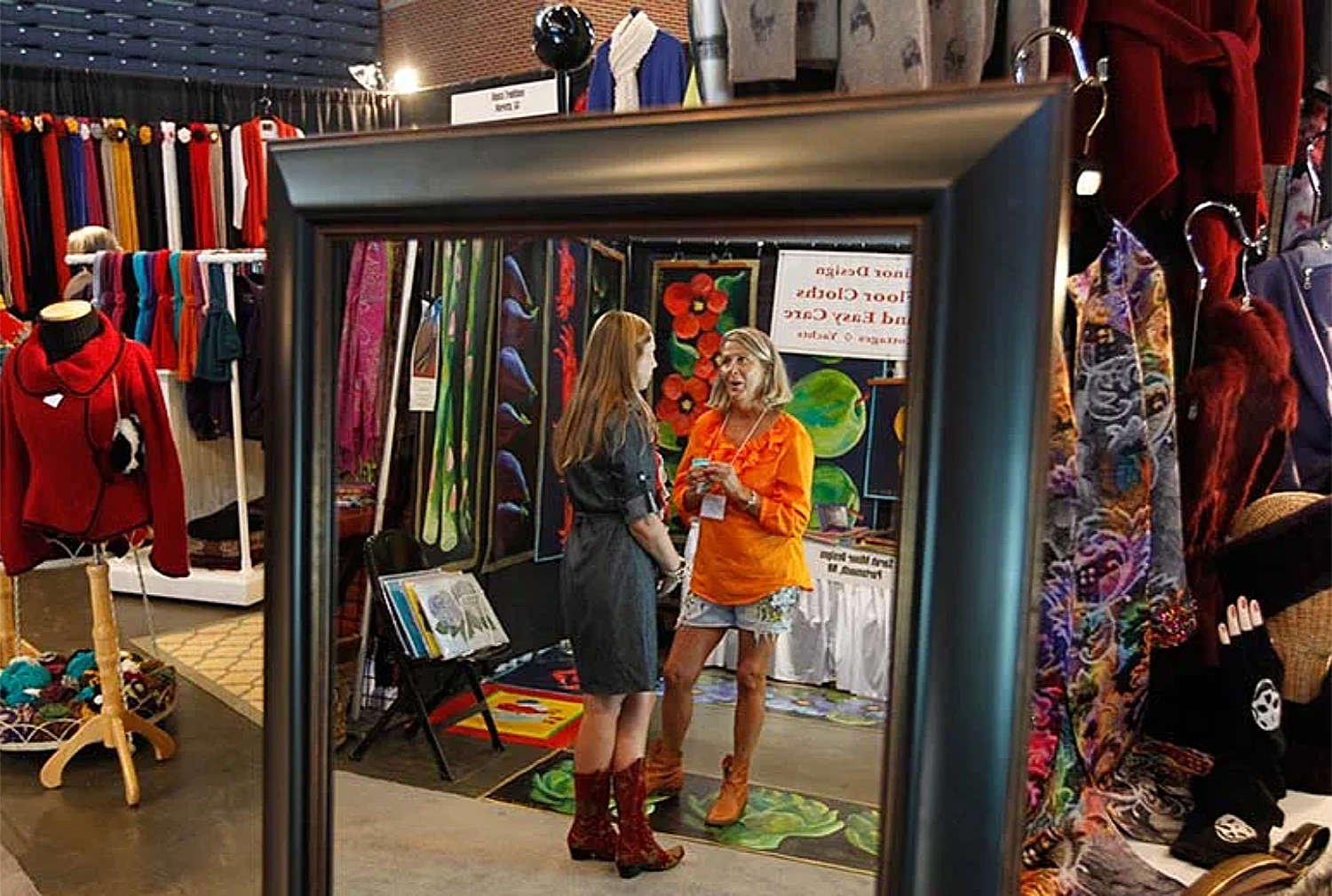 Two women talking at an indoor craft fair or market booth, with colorful textiles, clothing, and artwork on display around them, reflected in a large mirror.