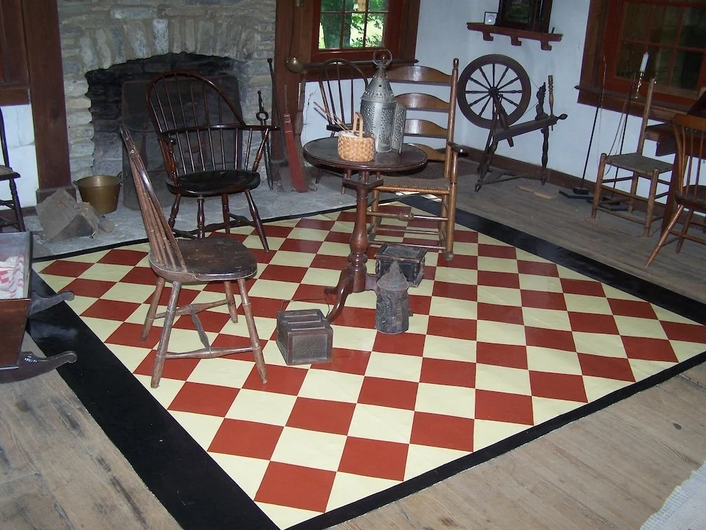 Indoor rustic room with stone fireplace, wooden chairs, spinning wheel, and checkerboard-patterned floor.