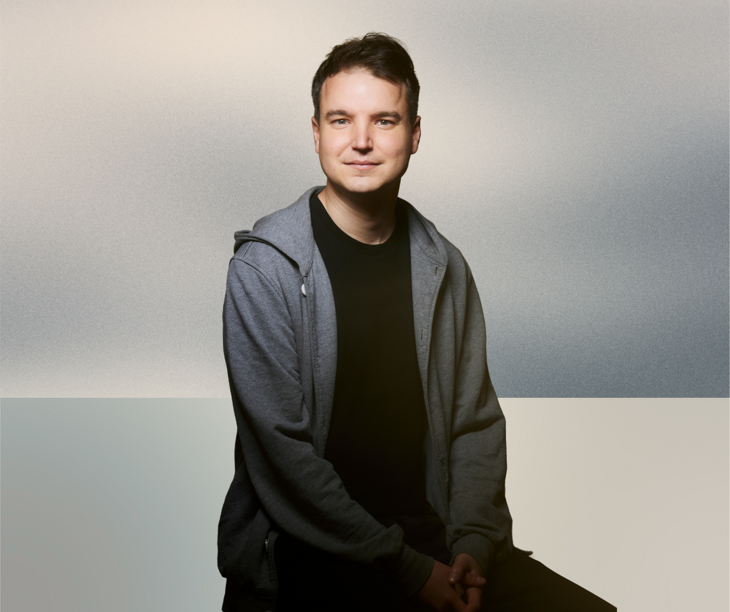 Porträt eines jungen Mannes mit dunkelbraunen Haaren, blauen Augen und leichtem Lächeln, trägt einen schwarzen T-Shirt und eine graue Jacke, vor einem hellgrauen Hintergrund.