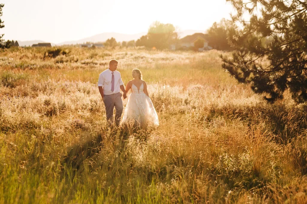 A couple in wedding attire walking hand in hand through a field at sunset.