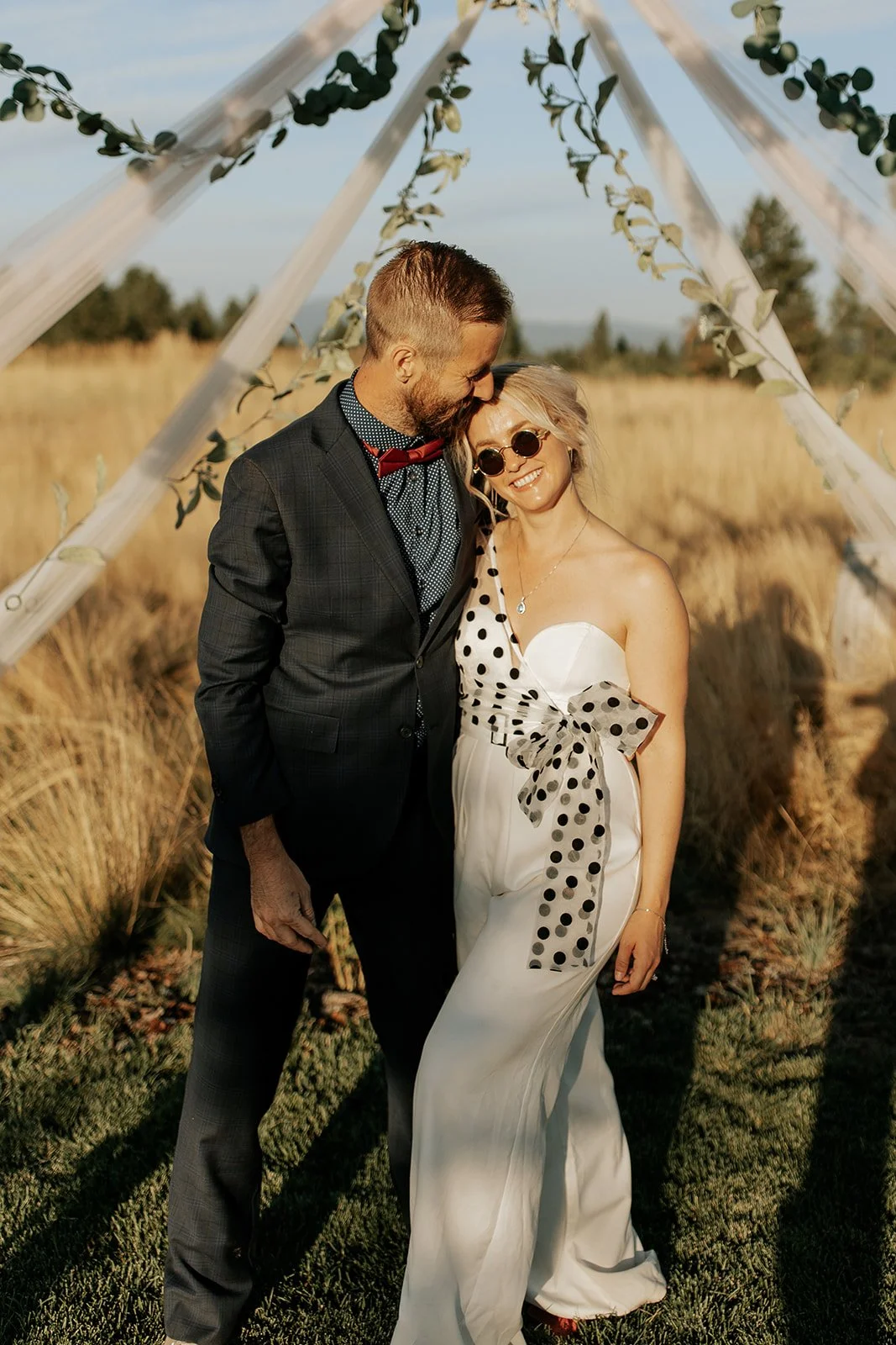 A couple in formal attire standing outside in a field, smiling and embracing under a wooden structure decorated with greenery for what appears to be a wedding or special event.