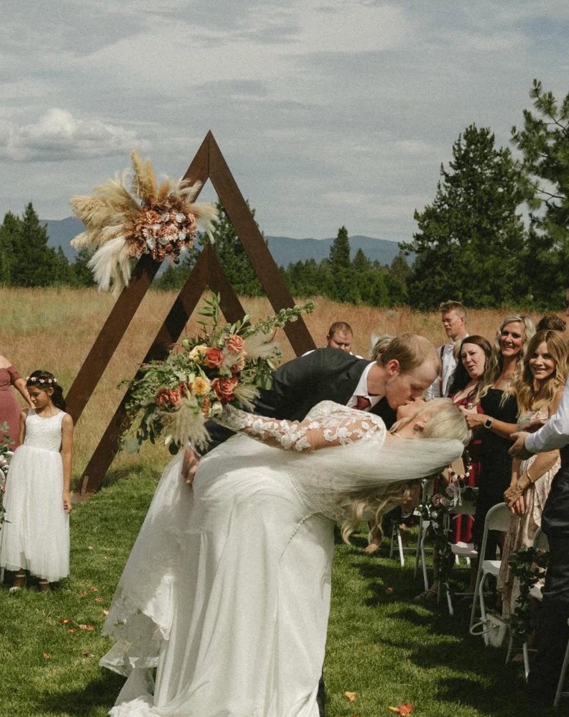 A wedding ceremony outdoors with a bride and groom kissing, surrounded by guests, with a decorative wooden triangle arch and outdoor natural scenery in the background.