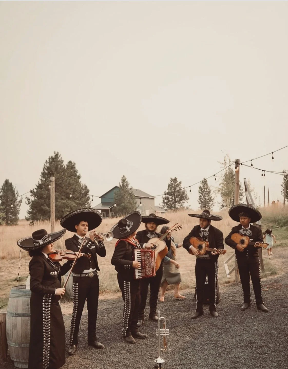 A group of six mariachi musicians wearing traditional clothing and sombreros playing instruments outdoors. The setting appears to be a rural area with trees, a house, and string lights in the background.