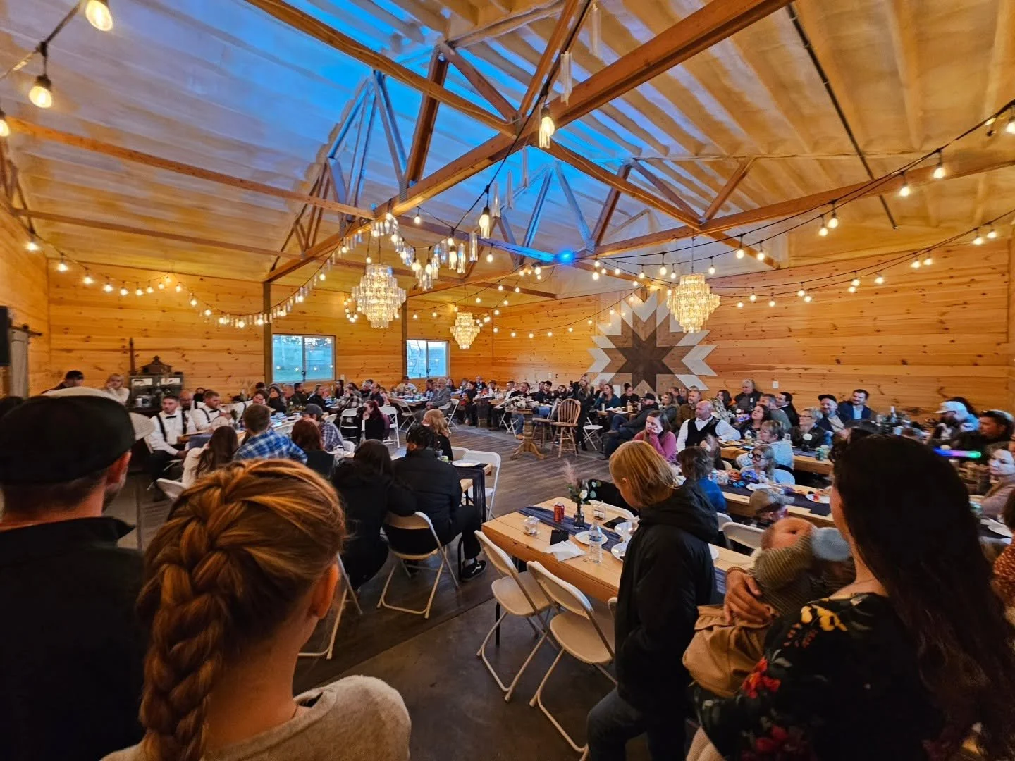 A large gathering of people sitting at tables in a decorated wooden hall for an event, with string lights and chandeliers hanging from the ceiling.