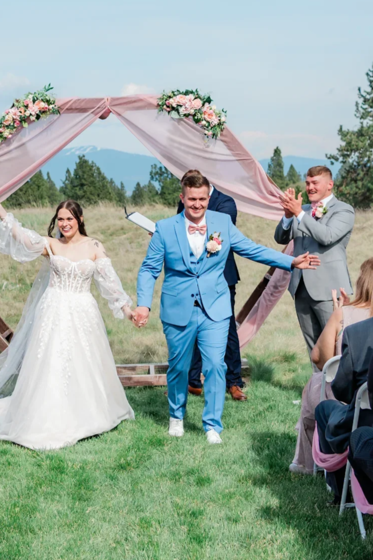 A newlywed couple holding hands and smiling at an outdoor wedding ceremony, with guests clapping and a floral arch in a scenic natural setting.