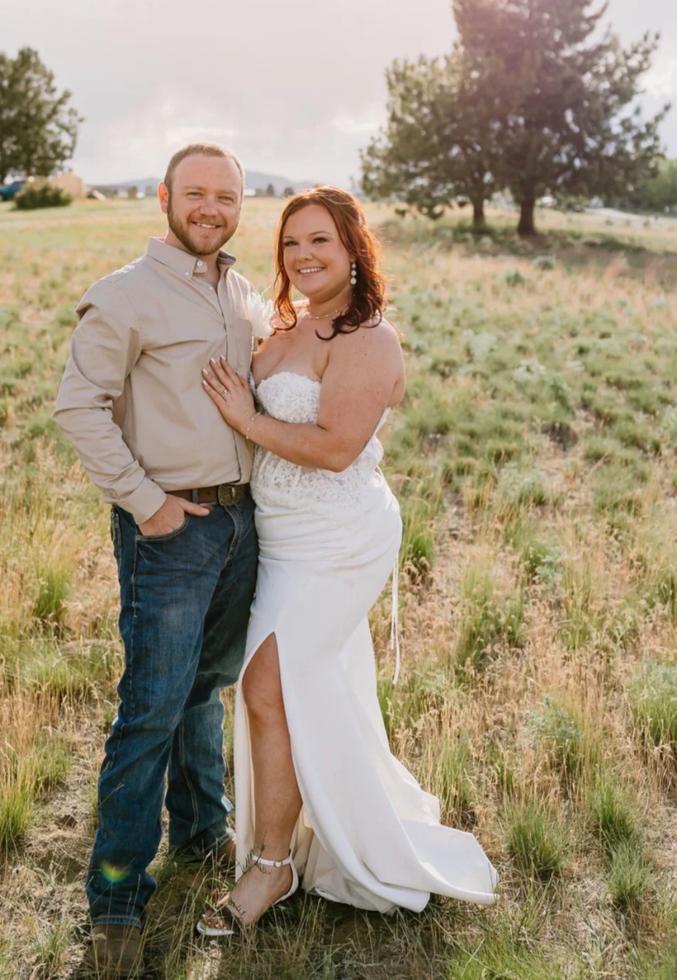 A couple in wedding attire standing in a grassy field during sunset, smiling. The woman is in a white strapless wedding dress with a slit, and the man is in a beige shirt and blue jeans.