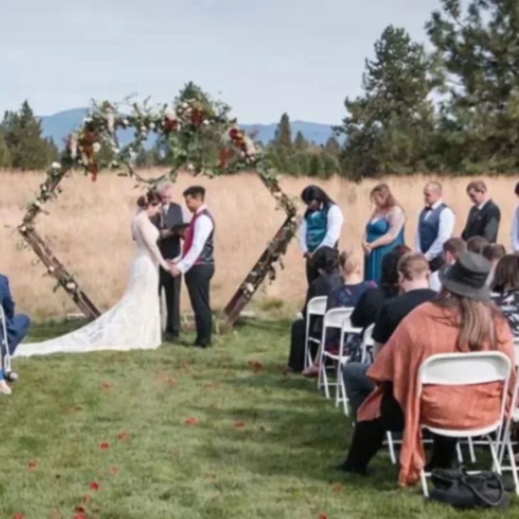A wedding ceremony outdoors with a couple exchanging vows under a floral arch in front of seated guests, with a backdrop of trees and mountains.