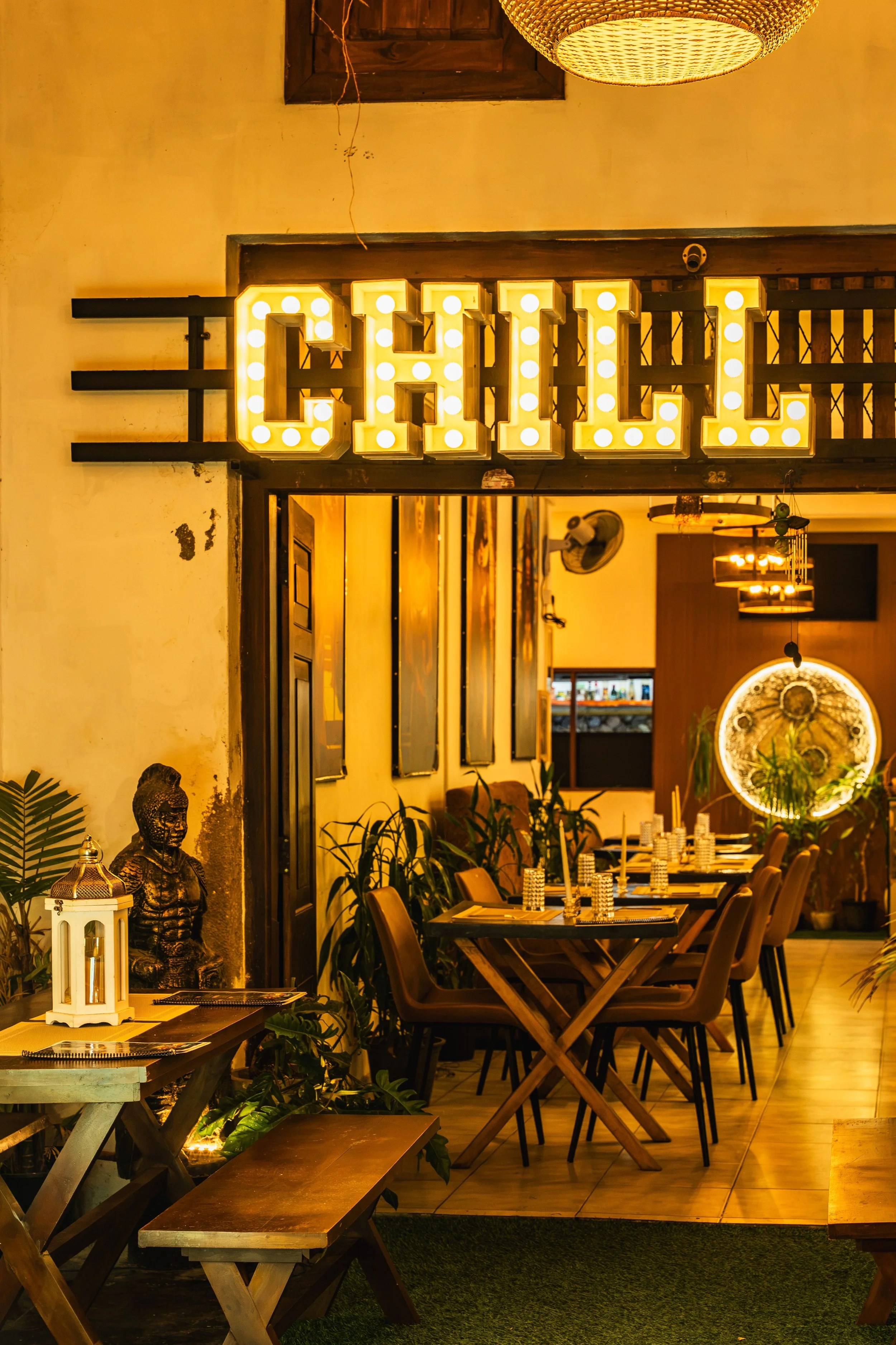 Interior de un restaurante con decoración cálida, mesas con manteles y vasos apilados, plantas y una estatua de Buda, iluminación tenue y un letrero brillante que dice 'COFFEE'.