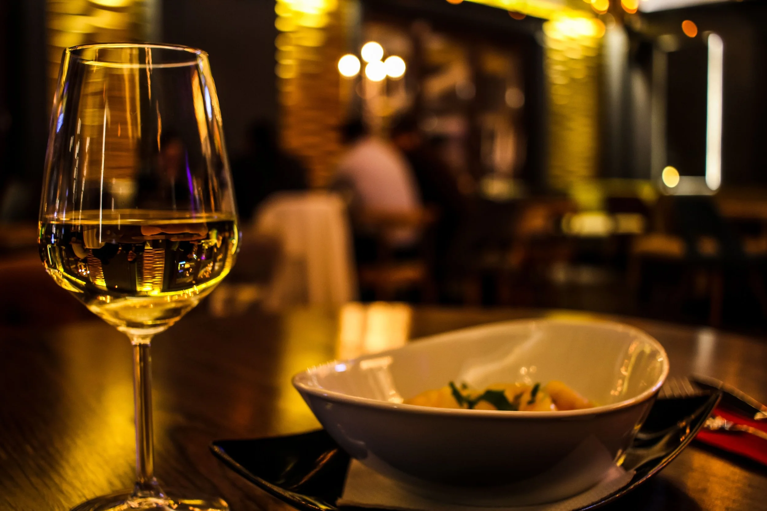 Copa de vino blanco en primer plano y un plato de comida en el fondo en un ambiente de restaurante con iluminación cálida.