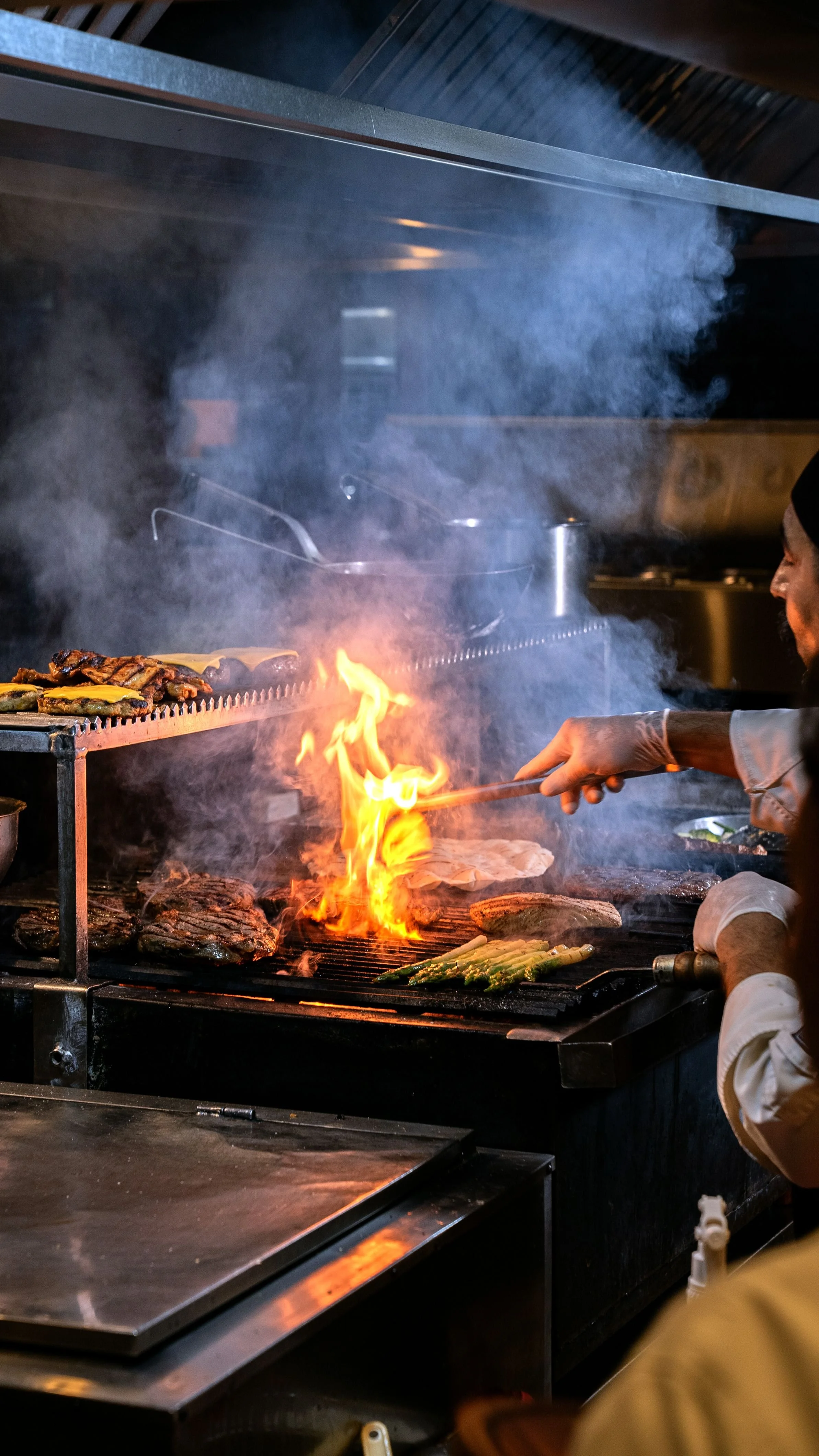 Un cocinero cocina carnes y verduras en una parrilla con llamas y humo.