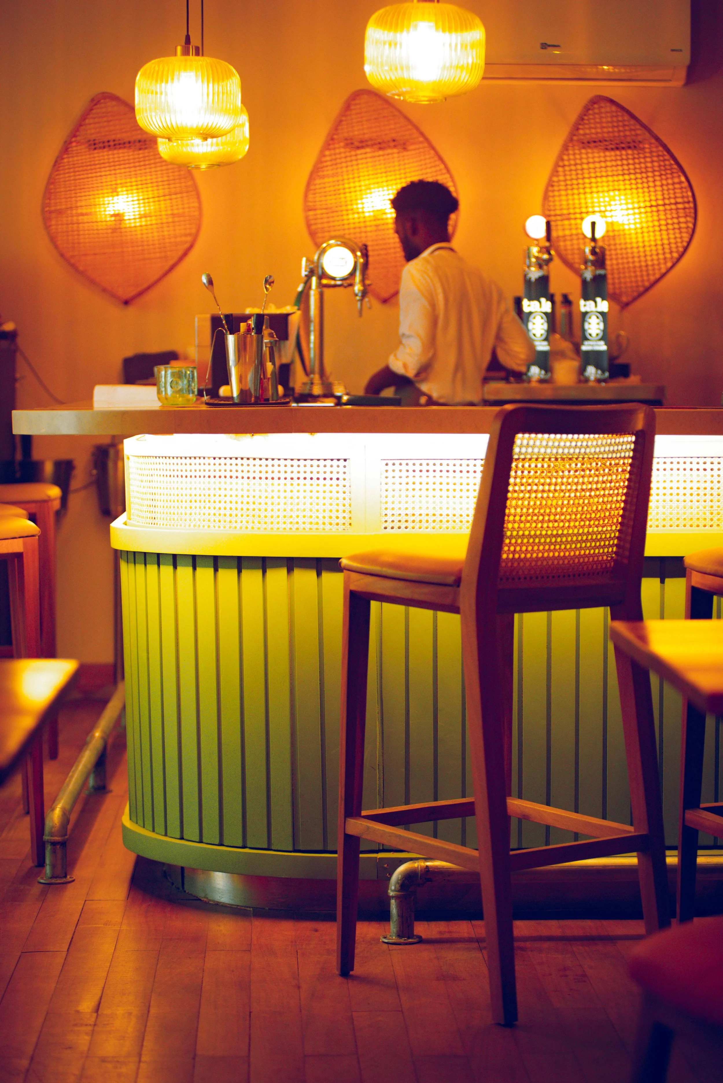 Interior de un bar con iluminación cálida, una barra de color verde con lamparitas doradas colgantes, un bartender sirviendo bebidas, varias sillas de madera alrededor y paredes decoradas con paneles de mimbre