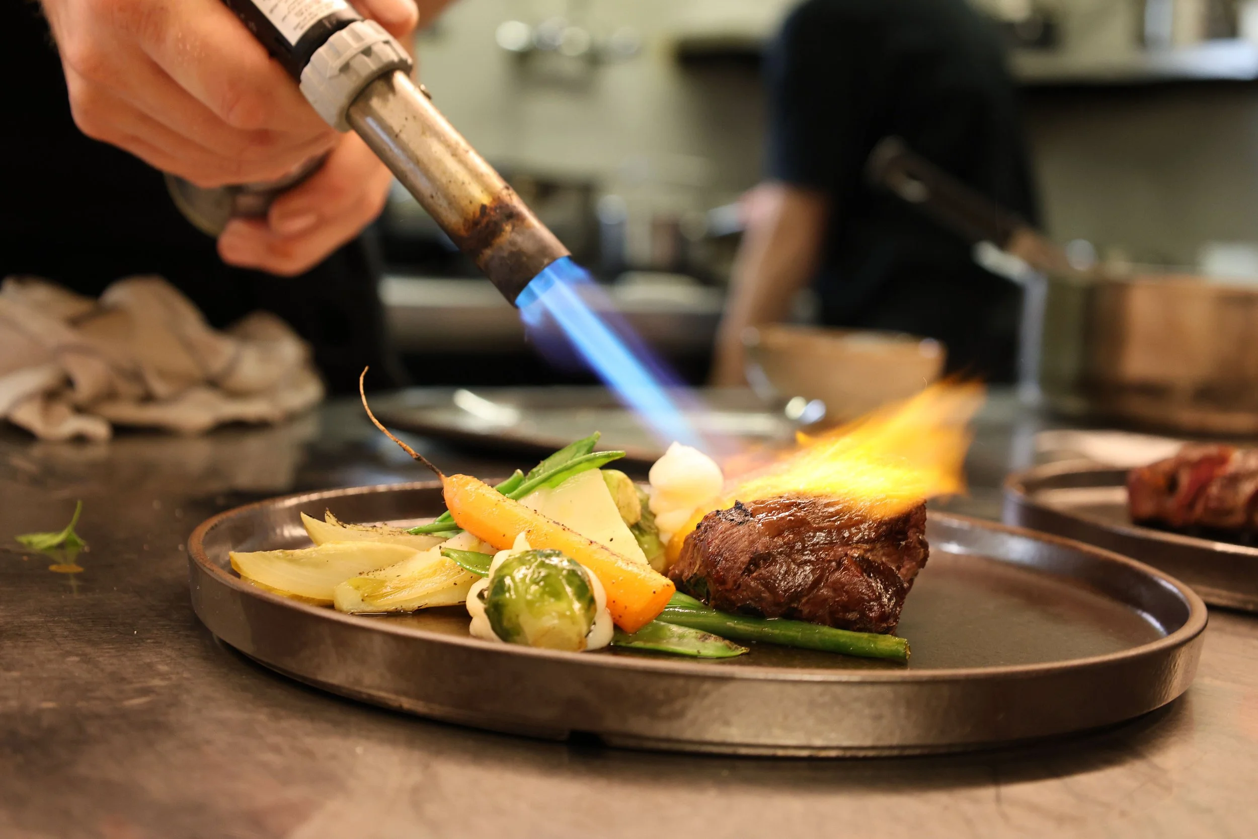Persona usando una antorcha para cocinar carne en un plato con verduras, en un ambiente de restaurante.