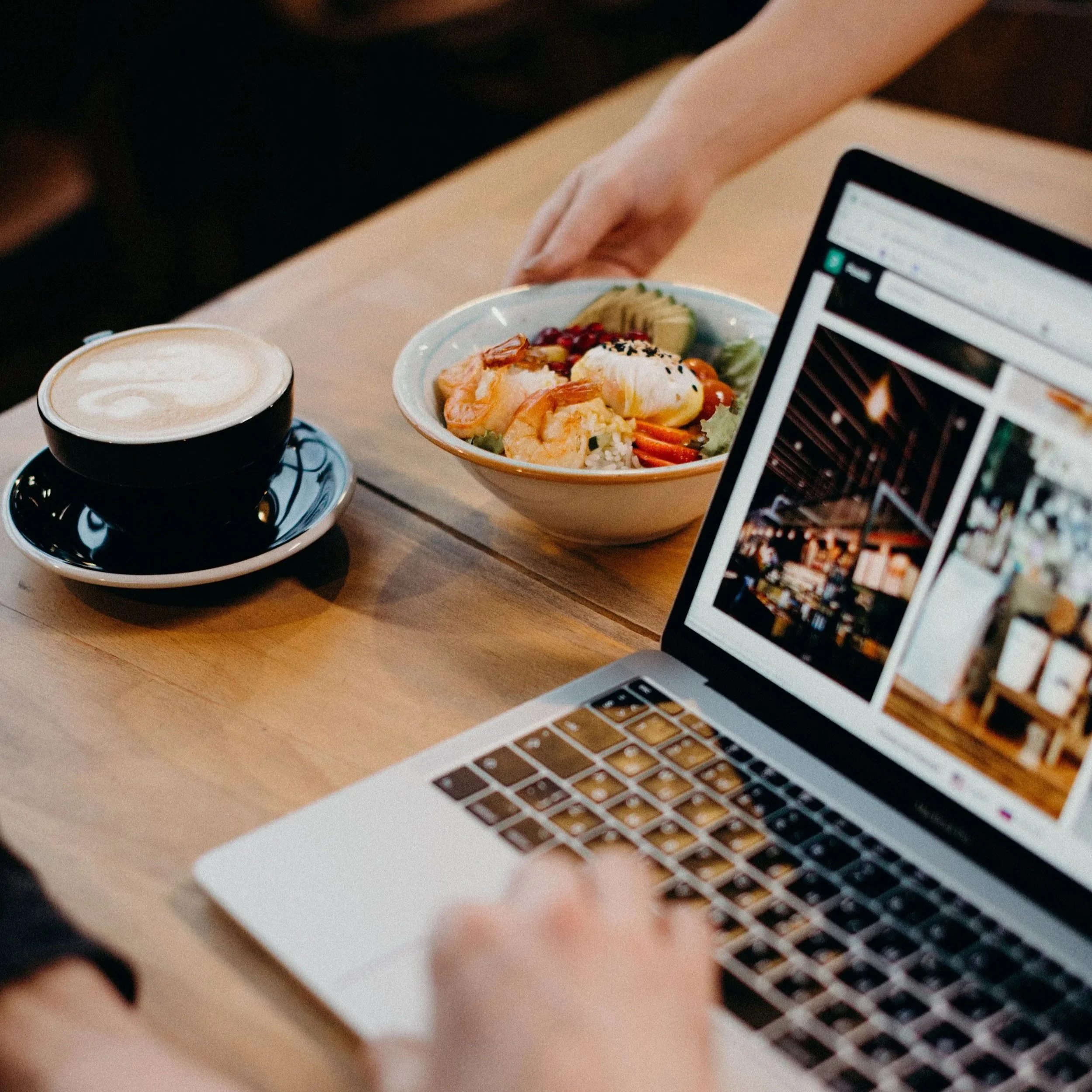 Una computadora portátil y una taza de café con leche en un escritorio. También hay un plato con comida y un teléfono móvil.