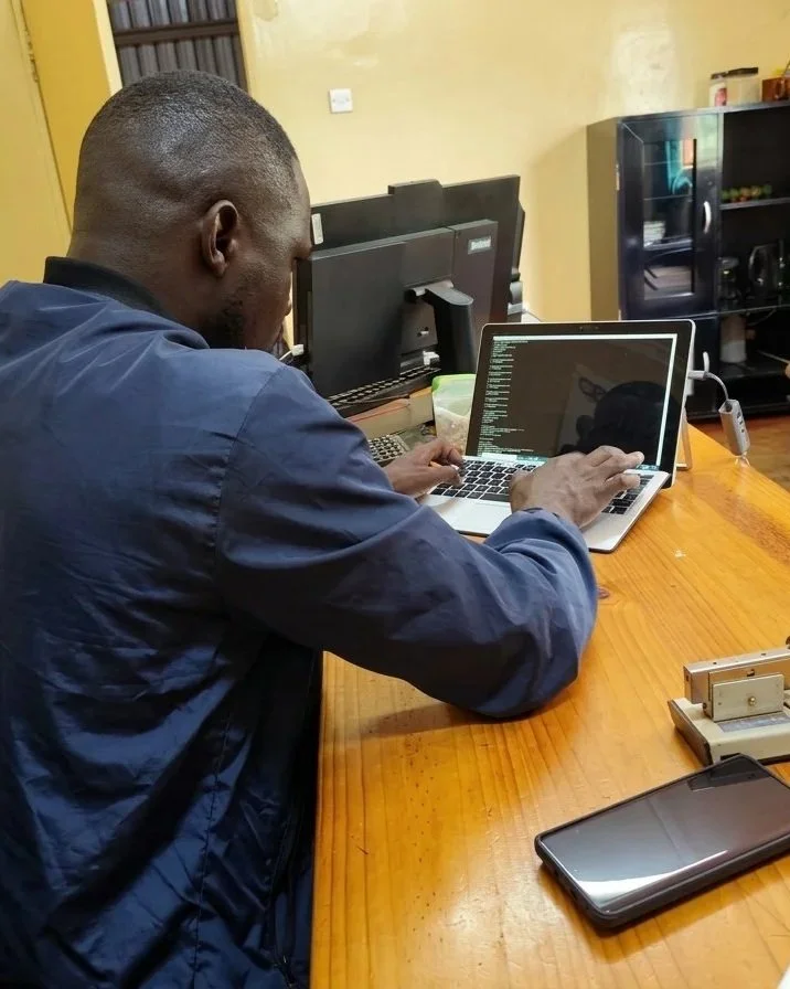A person working on a laptop at a wooden desk, with a smartphone and computer monitor nearby. The person is typing and appears focused on coding or programming, with a room-like environment in the background.