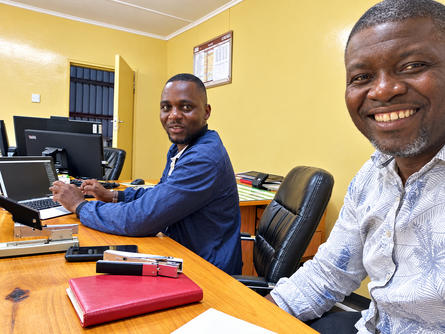 Two men smiling and sitting at a wooden conference table in an office, with computers, notebooks, and office supplies. The room has yellow walls, a door with a window, and a wall-mounted schedule or calendar.