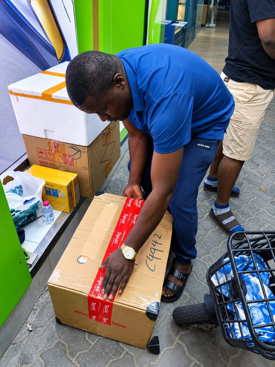 Man in blue shirt and sandals sealing a cardboard box with red tape outside a store, with shopping cart and other boxes nearby.