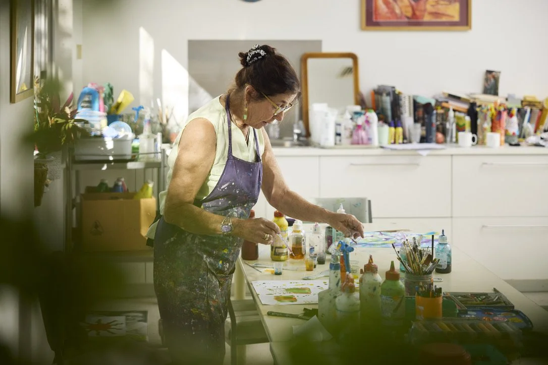 Dena working with paints and art materials at a sunlit studio table.