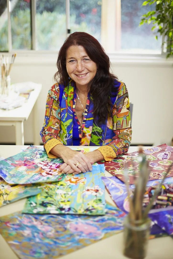 Dena Lawrence seated in her bright art studio, wearing a colourful patterned shirt.