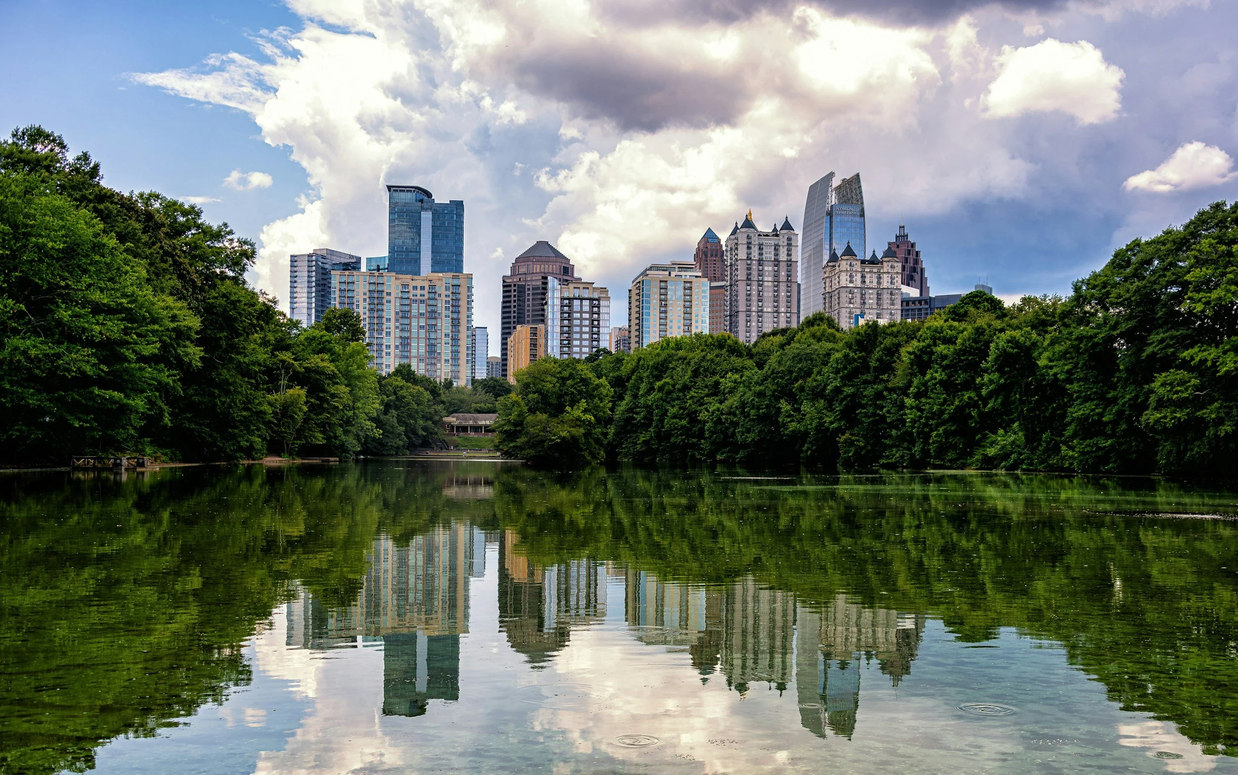 A city skyline with tall buildings reflecting in a river, and a lush green park with trees in the foreground, under a partly cloudy sky.