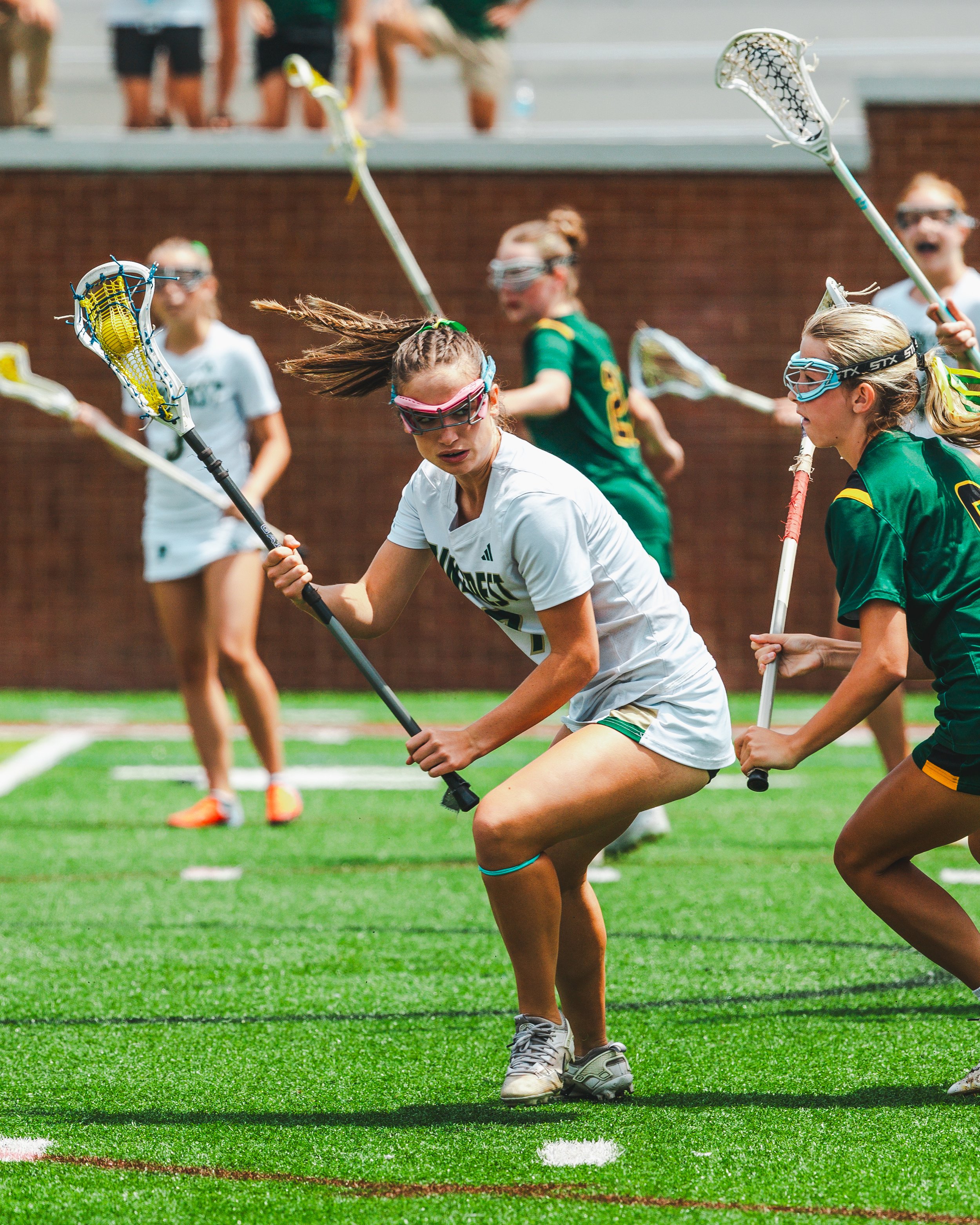 Youth girls playing lacrosse on a green field, wearing protective goggles and holding lacrosse sticks during a game.