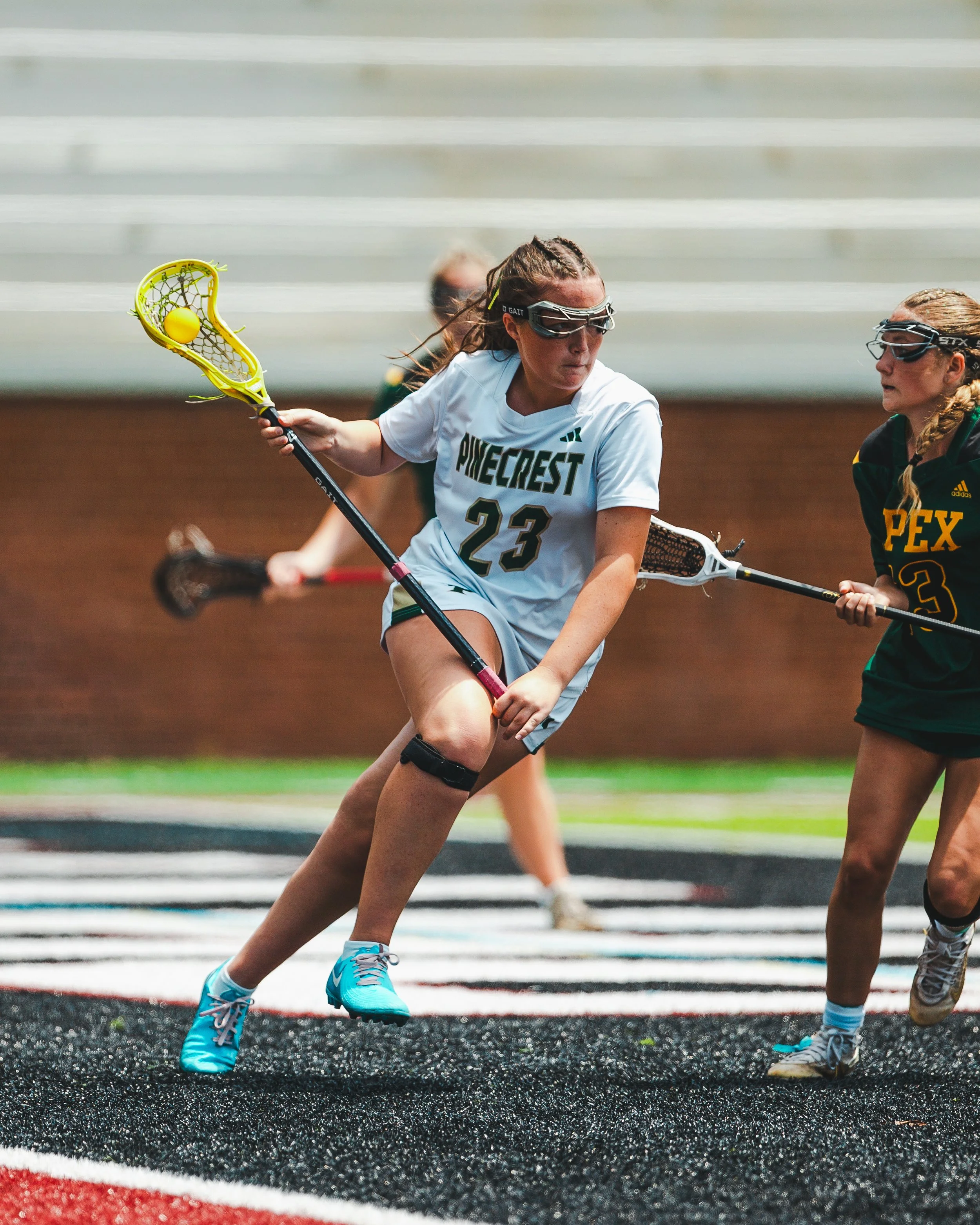 Two women playing lacrosse on a track field, one in a white Jersey with 'PINECREST' and the number 23, and the other in dark green with the letters 'PEX' and the number 23, both holding lacrosse sticks and wearing protective goggles.