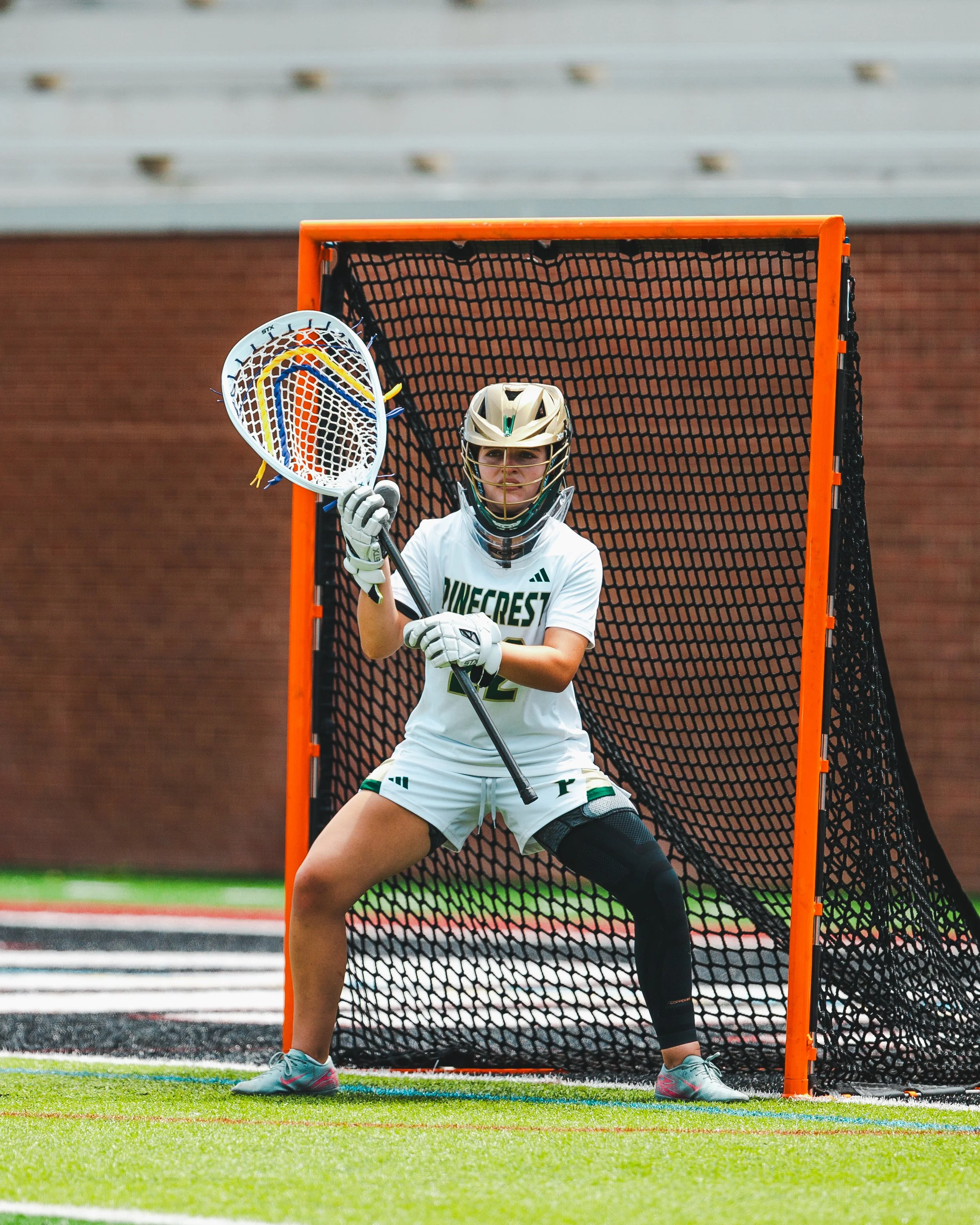 Lacrosse goalie stands in front of the goal, holding a lacrosse stick with a netted pocket, wearing protective gear including a helmet, gloves, and a jersey with the word 'GAMECROSS' on it, on a sports field.