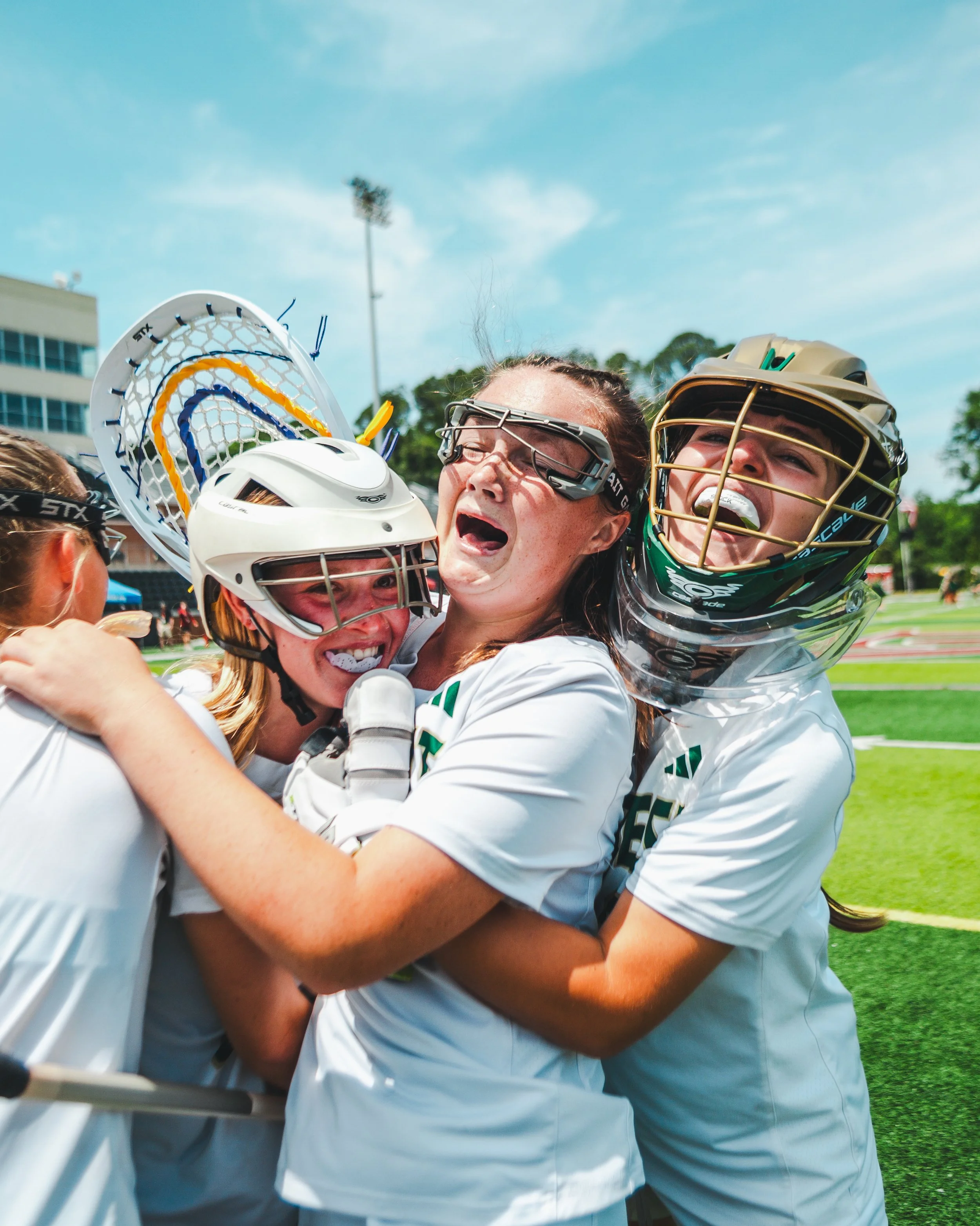 A group of female lacrosse players celebrating and hugging each other on a sports field during daytime, wearing helmets and sports uniforms.