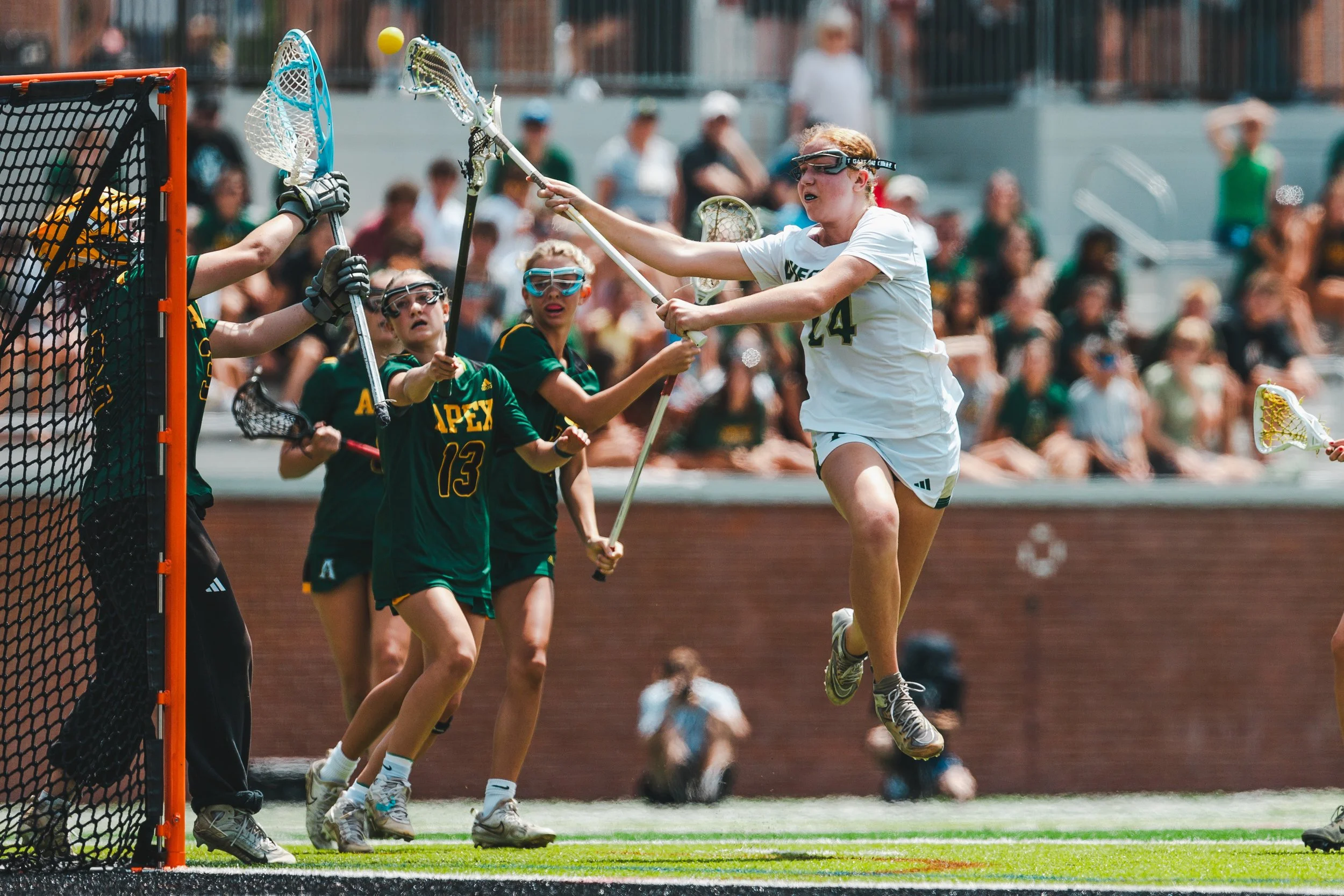 A female lacrosse player in a white uniform with the number 24 is jumping to block a shot near the goal, while other players and a goalie in green uniforms watch, during a game on a sunny day with spectators in the background.