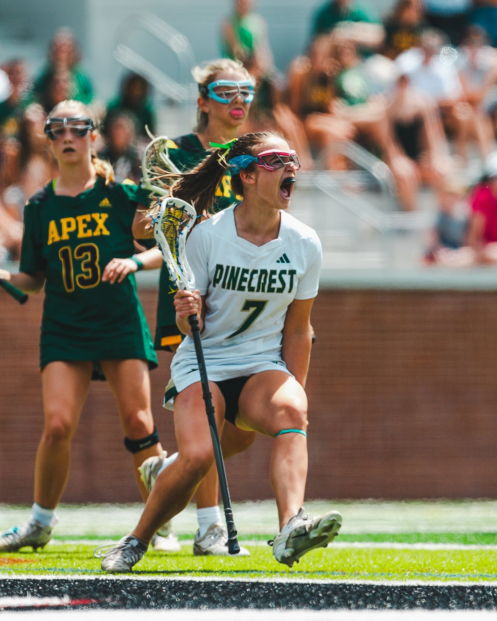 Women playing field hockey during a game, wearing uniforms, goggles, and holding hockey sticks, on a green turf field with audience in the background.