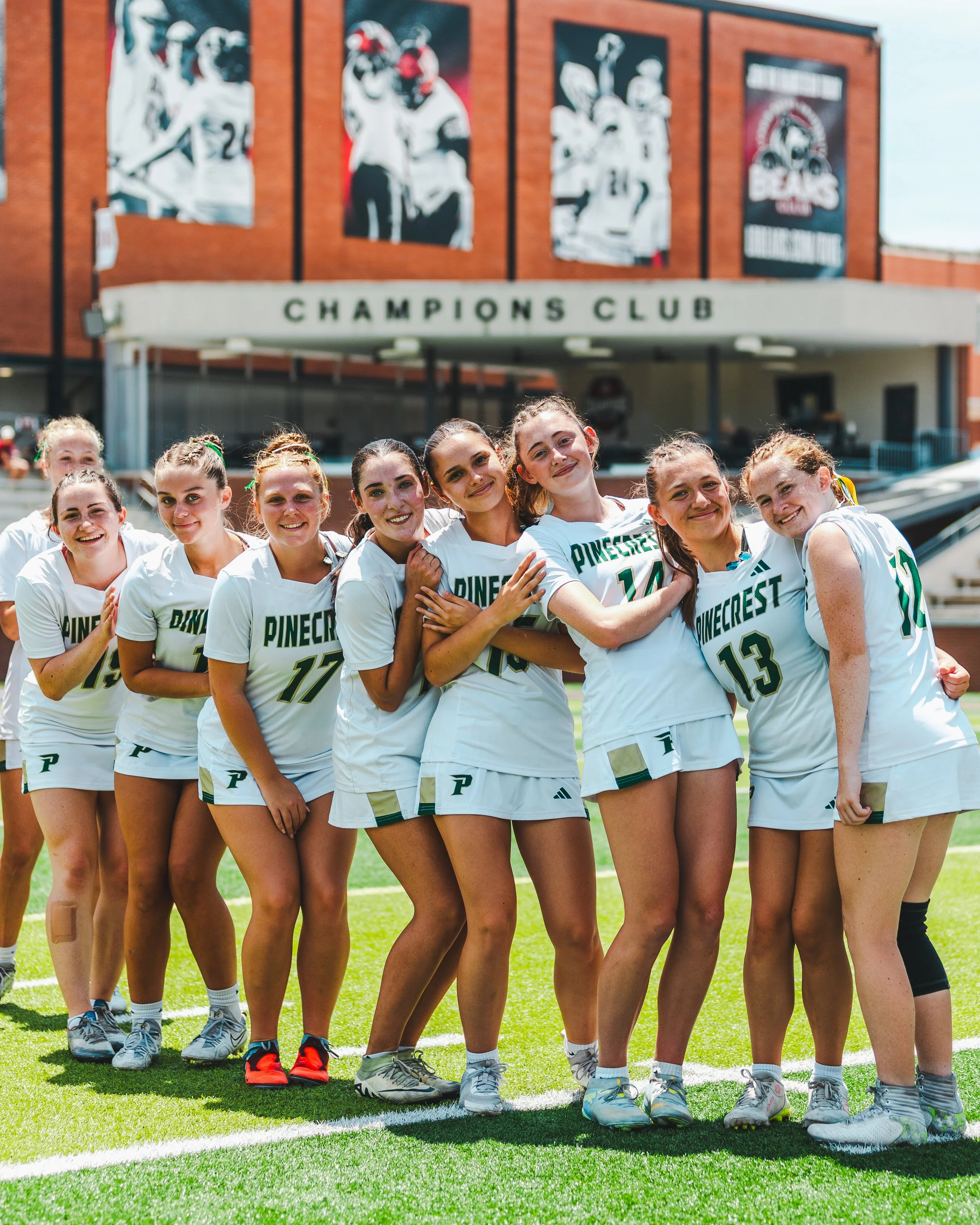 A group of young female athletes in white sport jerseys with green and gold lettering, standing close together on a sports field, smiling for a team photo.