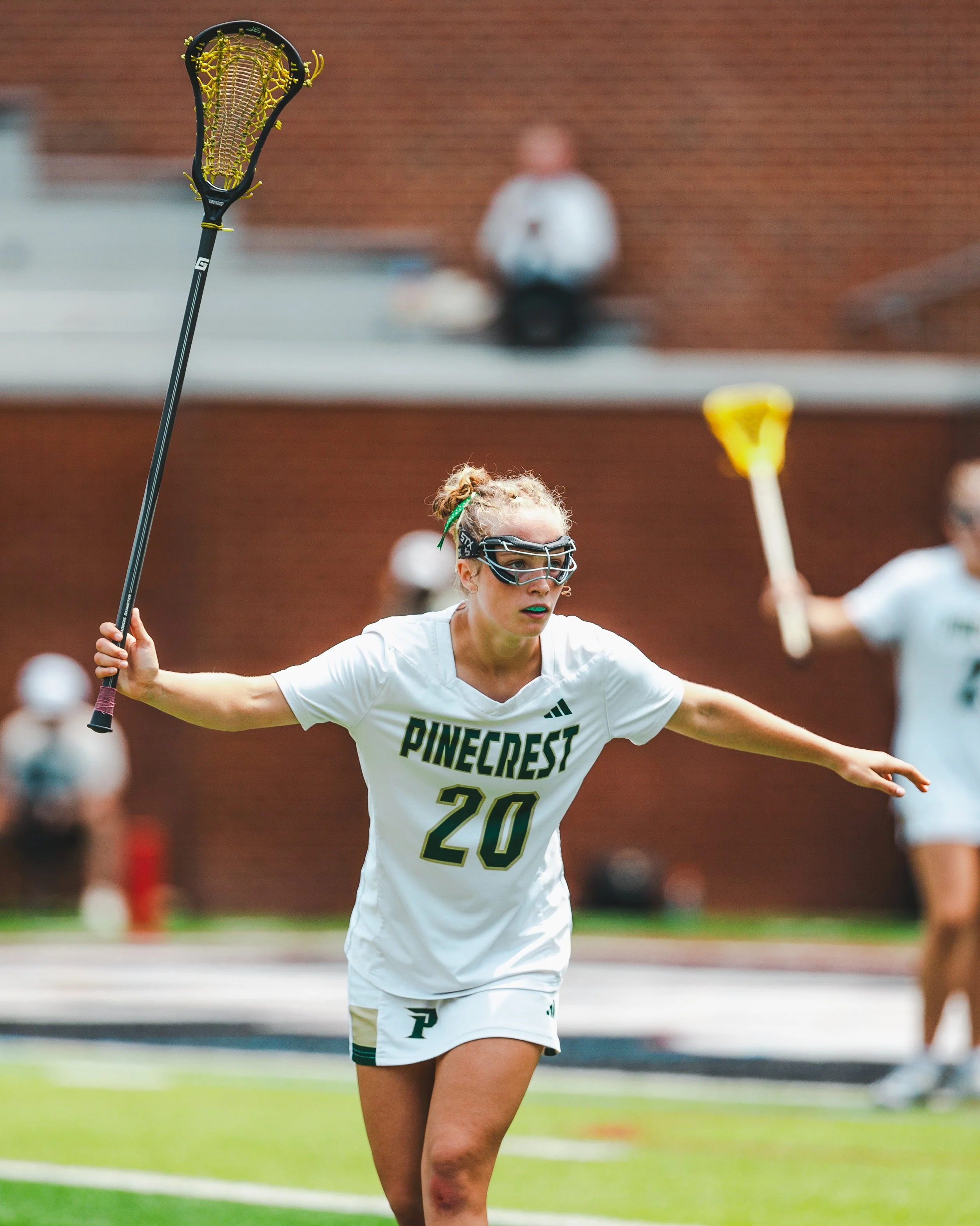 Female lacrosse player in white jersey with 'PINECREST' written on it, wearing sports goggles and holding a lacrosse stick, with her arm extended outward during a game or practice.