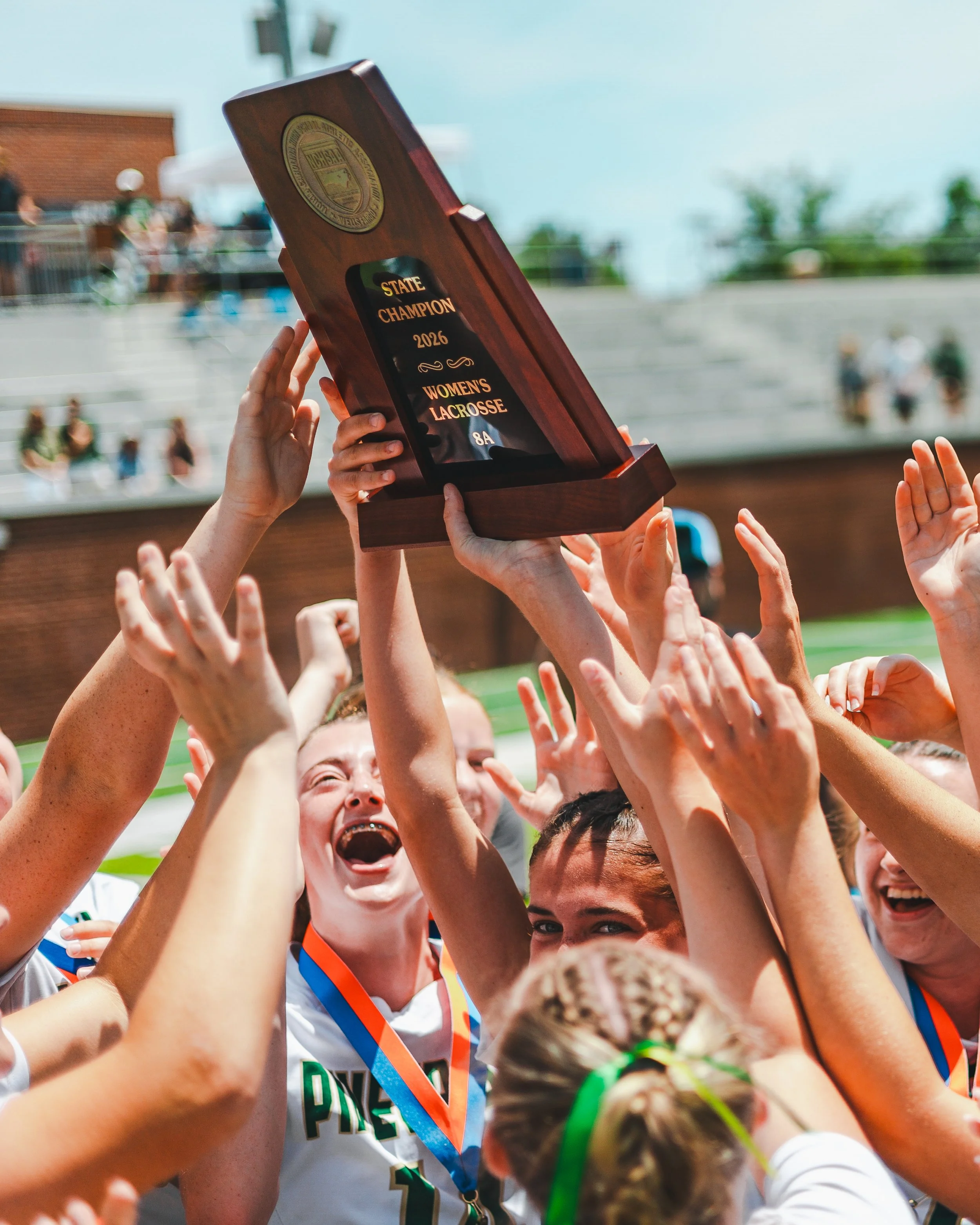 Group of people holding up a trophy that reads 'State Champion 2026 Women's Lacrosse 8A' during a celebration event.