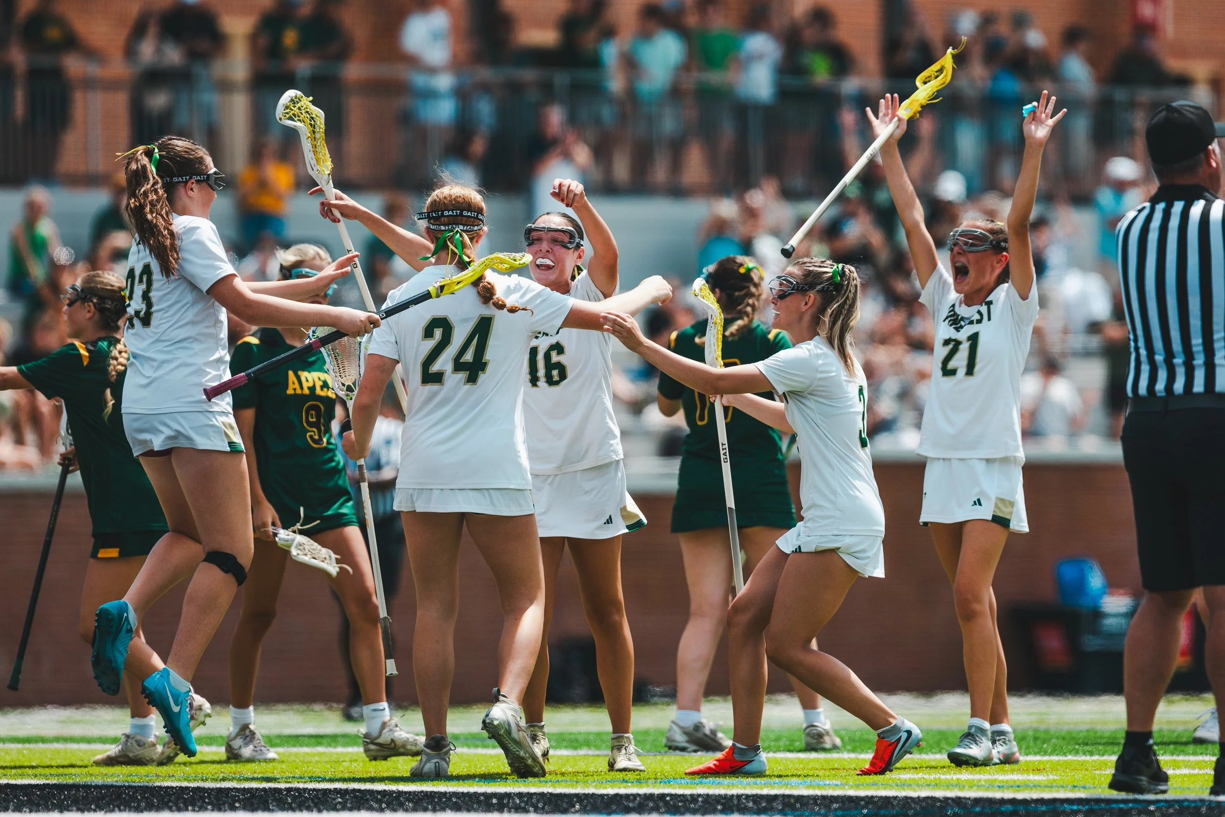 A group of girls playing lacrosse on a field, celebrating and high-fiving each other after a game or scoring, with a referee nearby and spectators in the background.