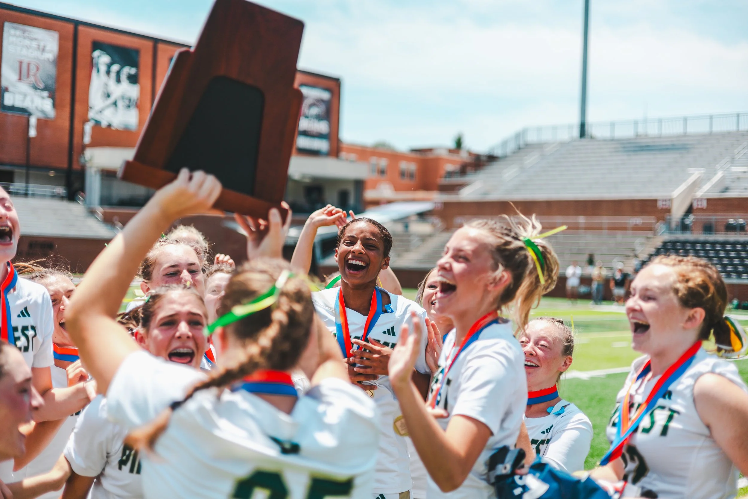 A group of young female soccer players celebrating on the field after winning a game, with some holding a trophy and wearing medals.
